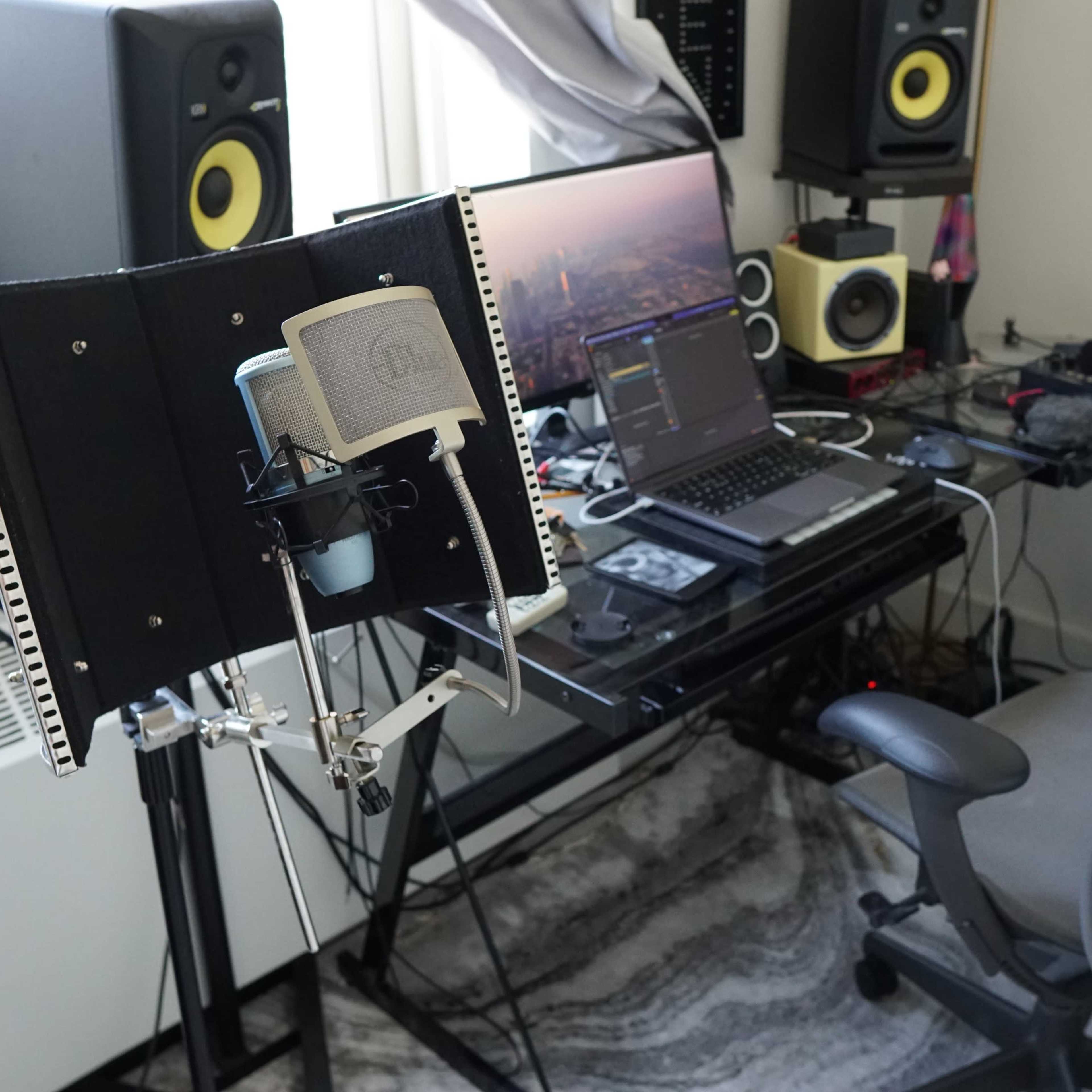A recording studio setup features a microphone on a stand, a computer with an audio interface, and various music equipment surrounded by cables and a desk chair.