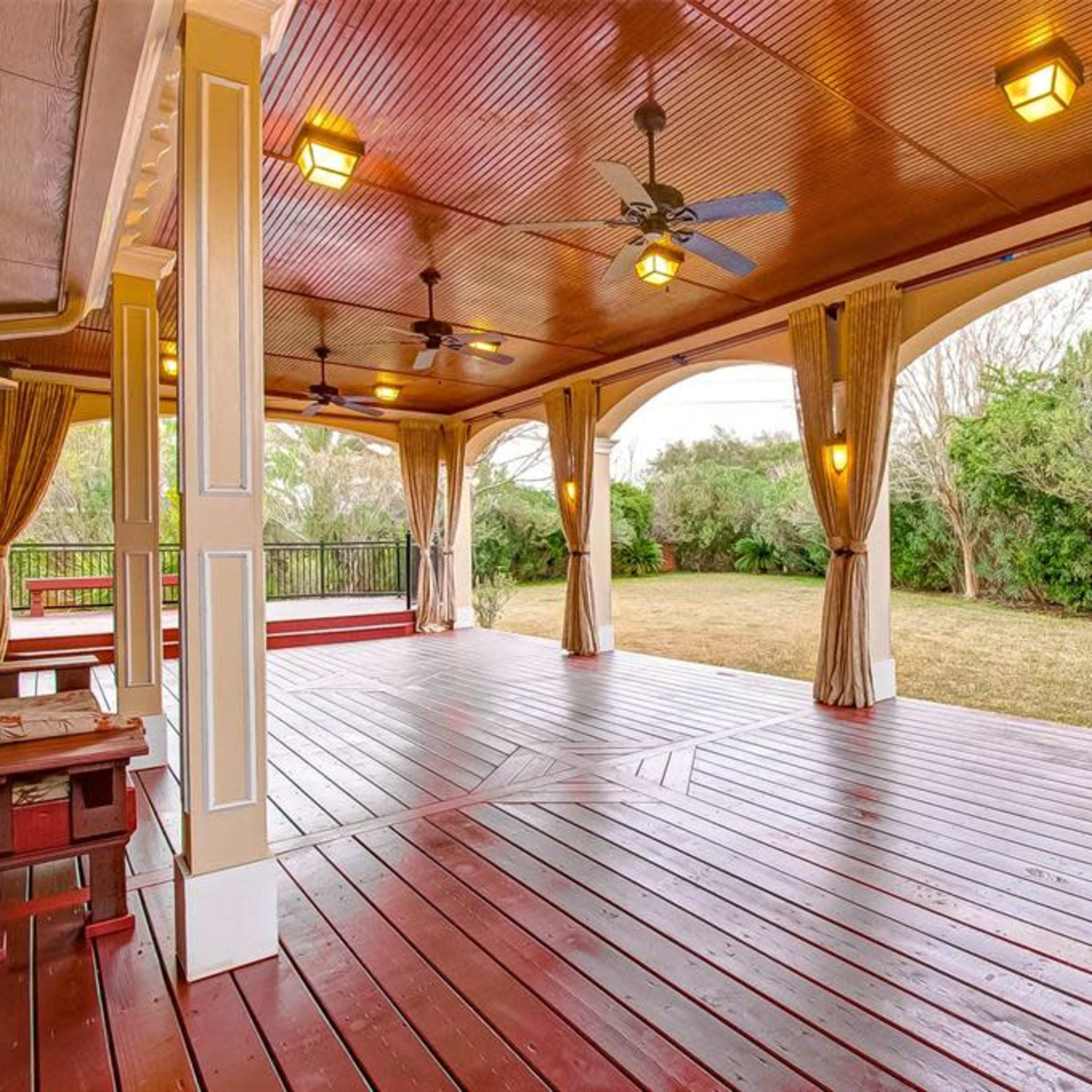 A large, covered patio with wooden flooring, ceiling fans, and curtains, overlooking a grassy backyard.