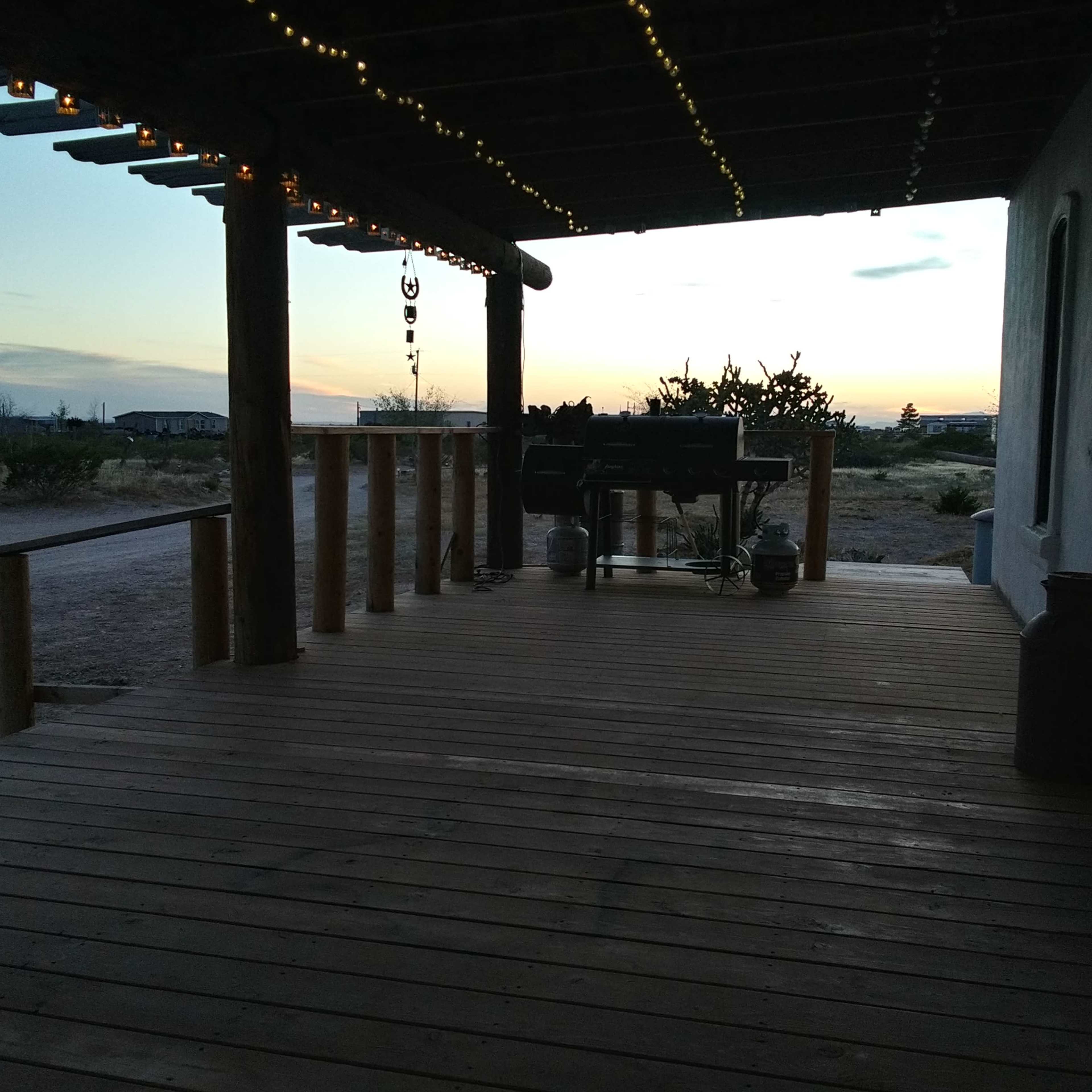 A wooden porch with string lights overlooks a dirt path and desert landscape under a colorful sky at dusk.
