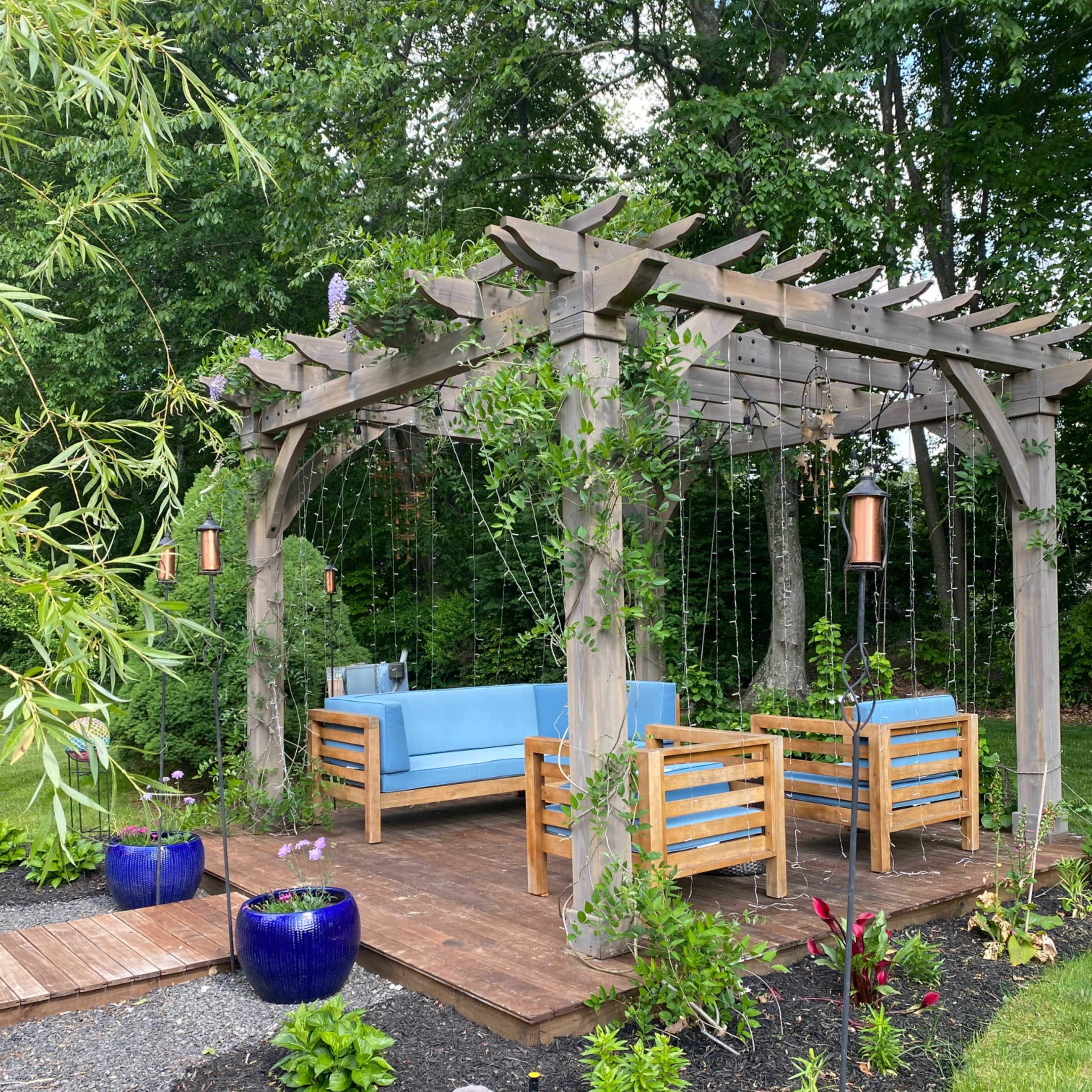 A wooden pergola with blue seating surrounded by greenery and decorative planters in a landscaped garden.