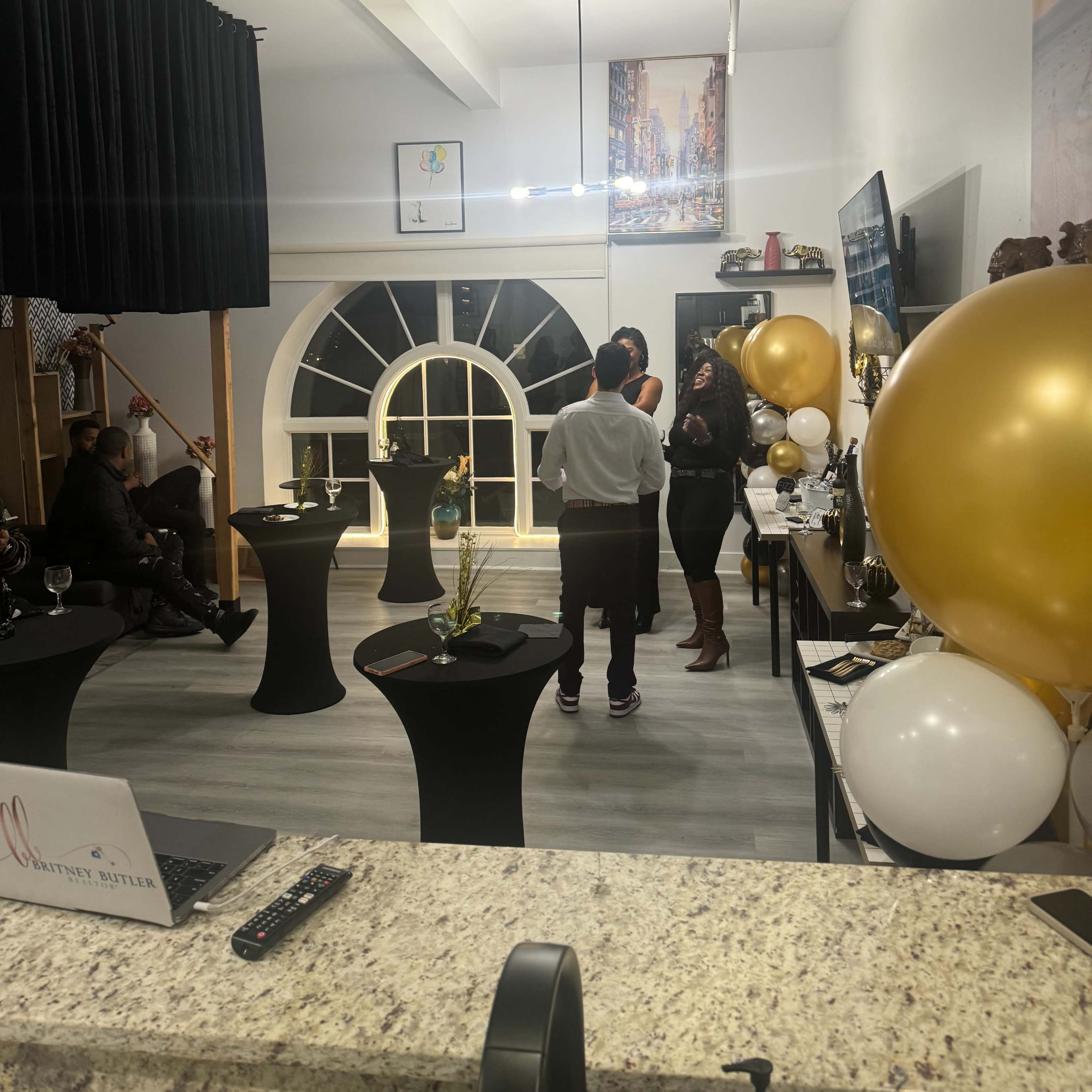 A cozy event space with a few guests conversing near elegantly arranged tables, decorated with black and gold balloons.