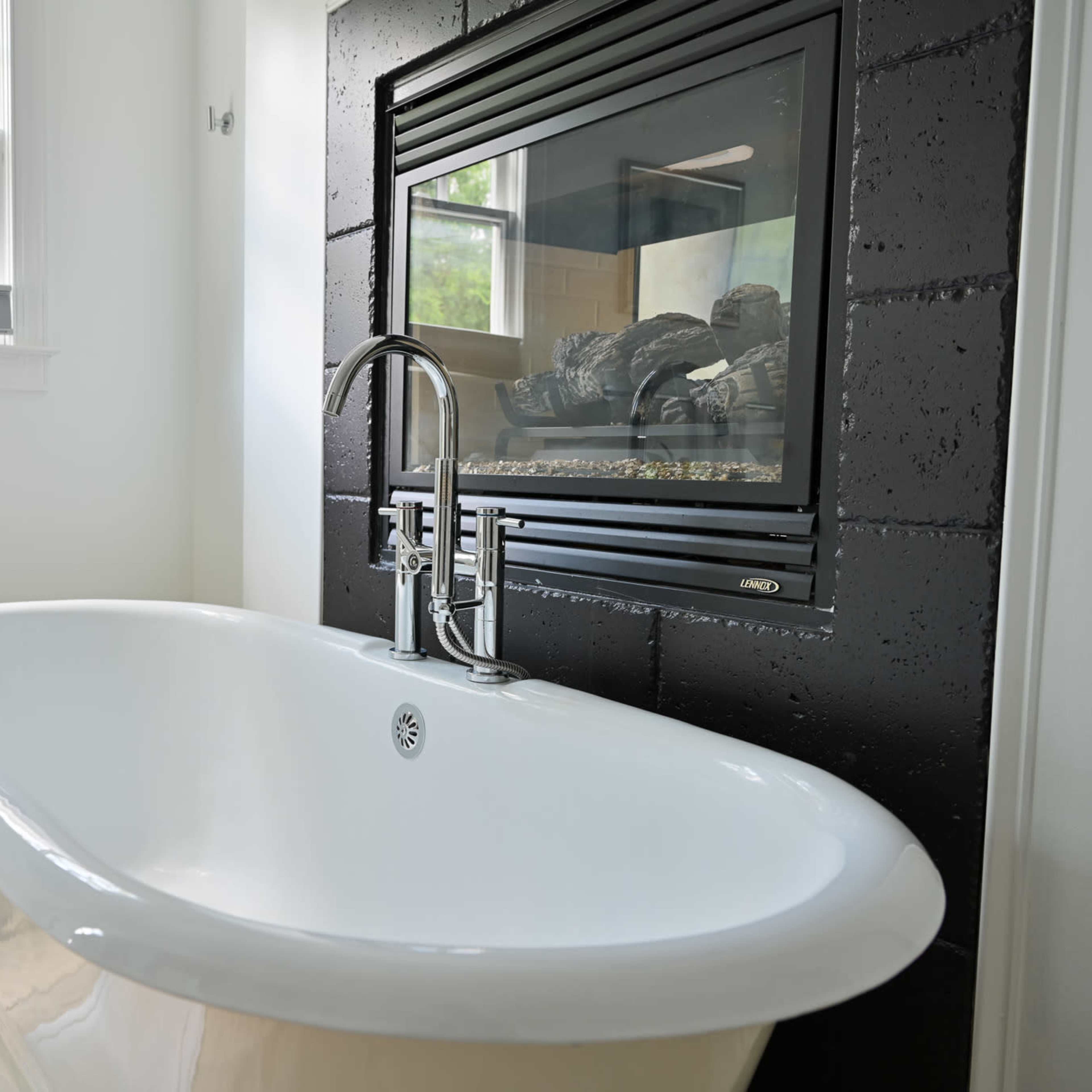 A modern bathroom with a freestanding white bathtub, a small table with a potted plant, and a fireplace embedded in a feature wall.