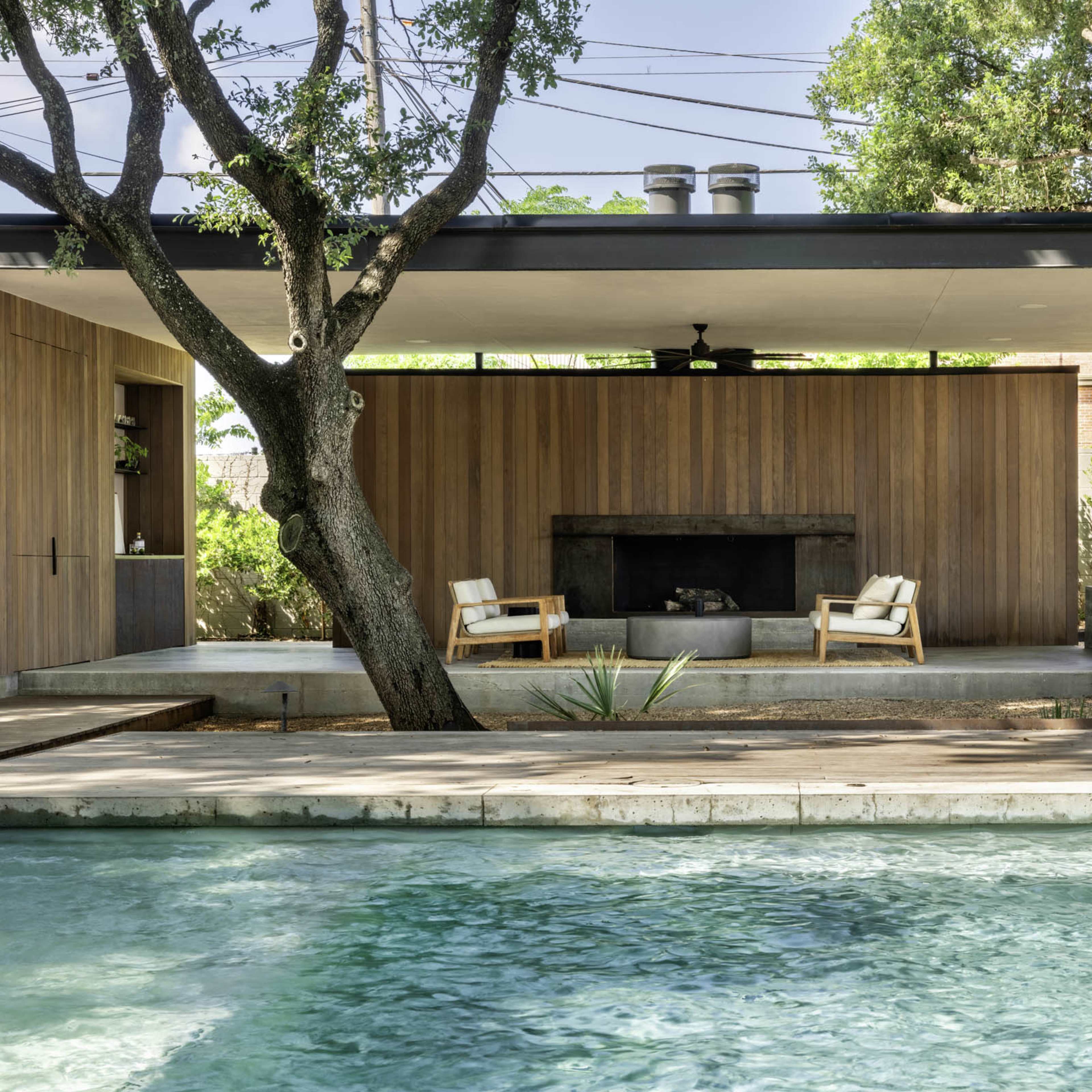A modern outdoor space features a pool, two chairs positioned by a fireplace, and a wooden structure surrounded by greenery.