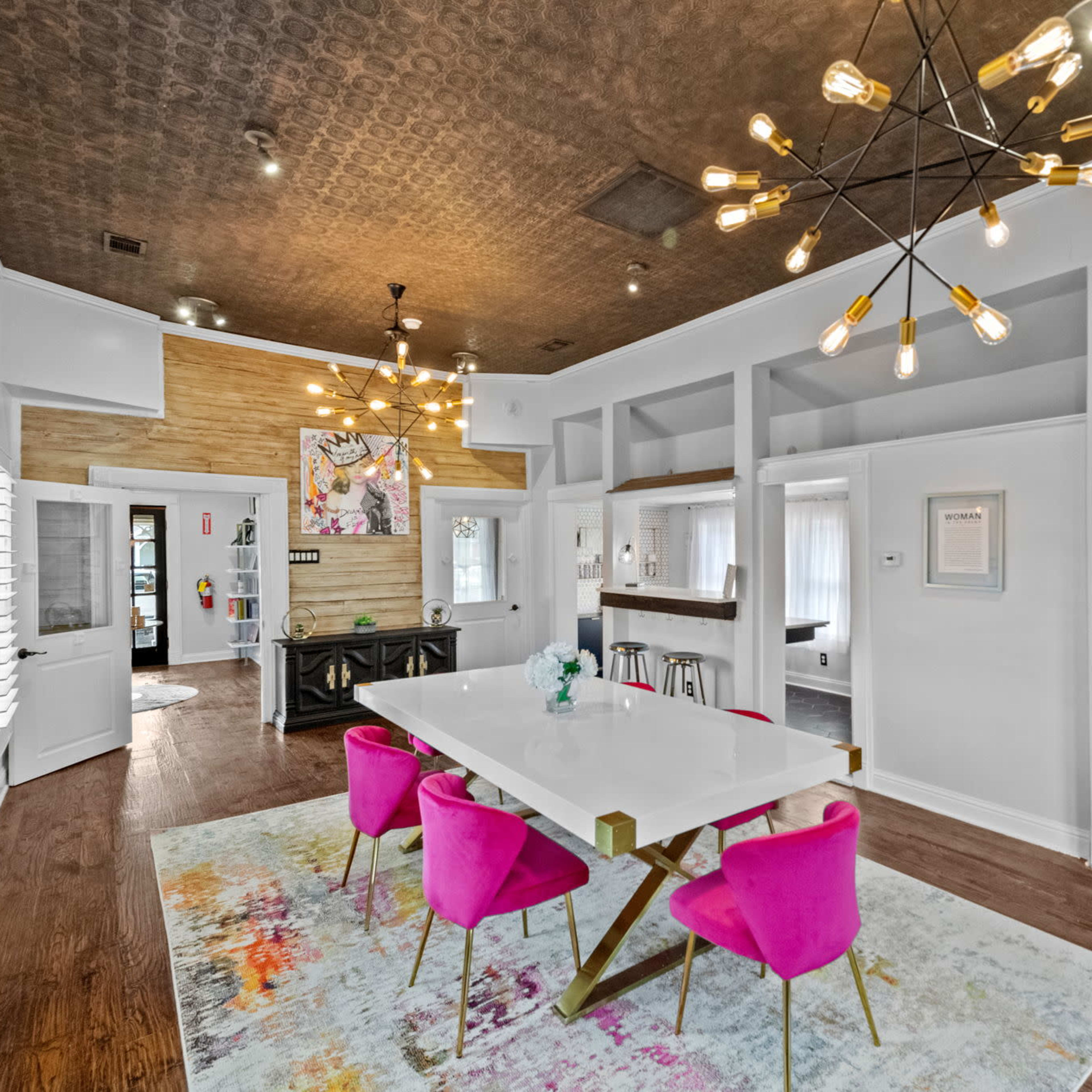 A modern interior space features a dining table surrounded by pink chairs, sleek lighting fixtures, and wooden accents on the walls.