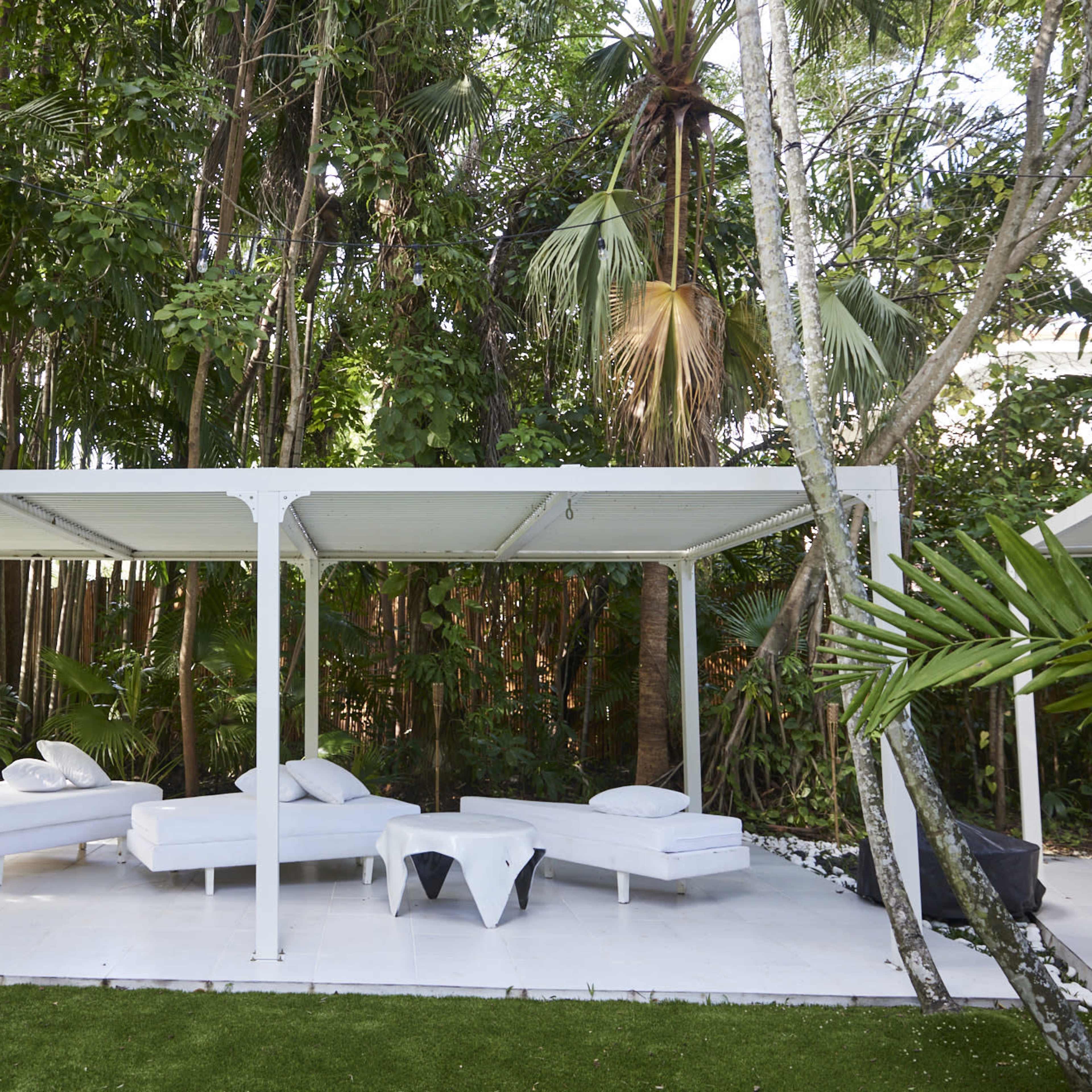 A white outdoor lounge area with several beds is situated under a pergola, surrounded by lush tropical vegetation.