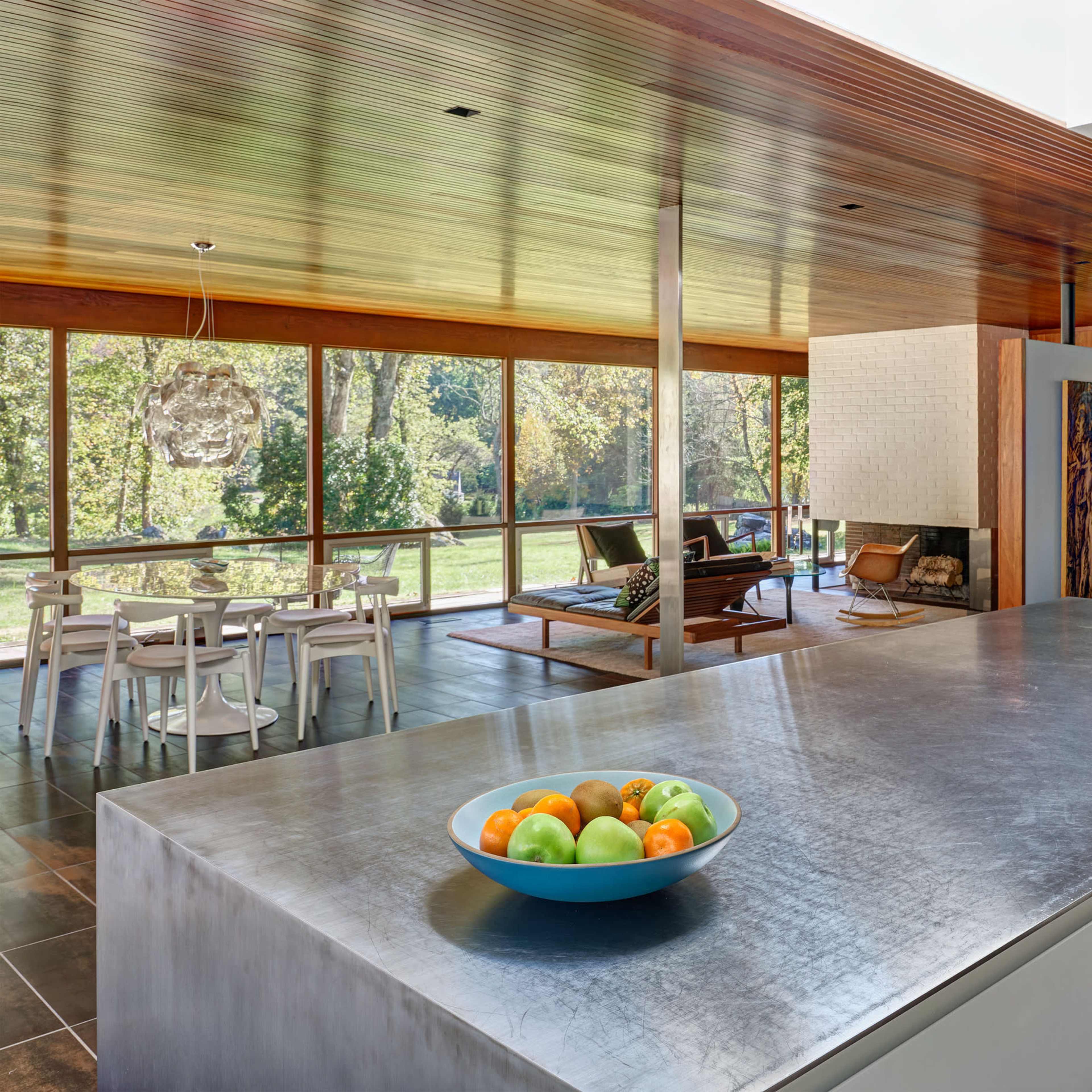 The kitchen features a stainless steel island with a bowl of fruit, overlooking a spacious dining area and large windows that reveal a view of the outdoor landscape.