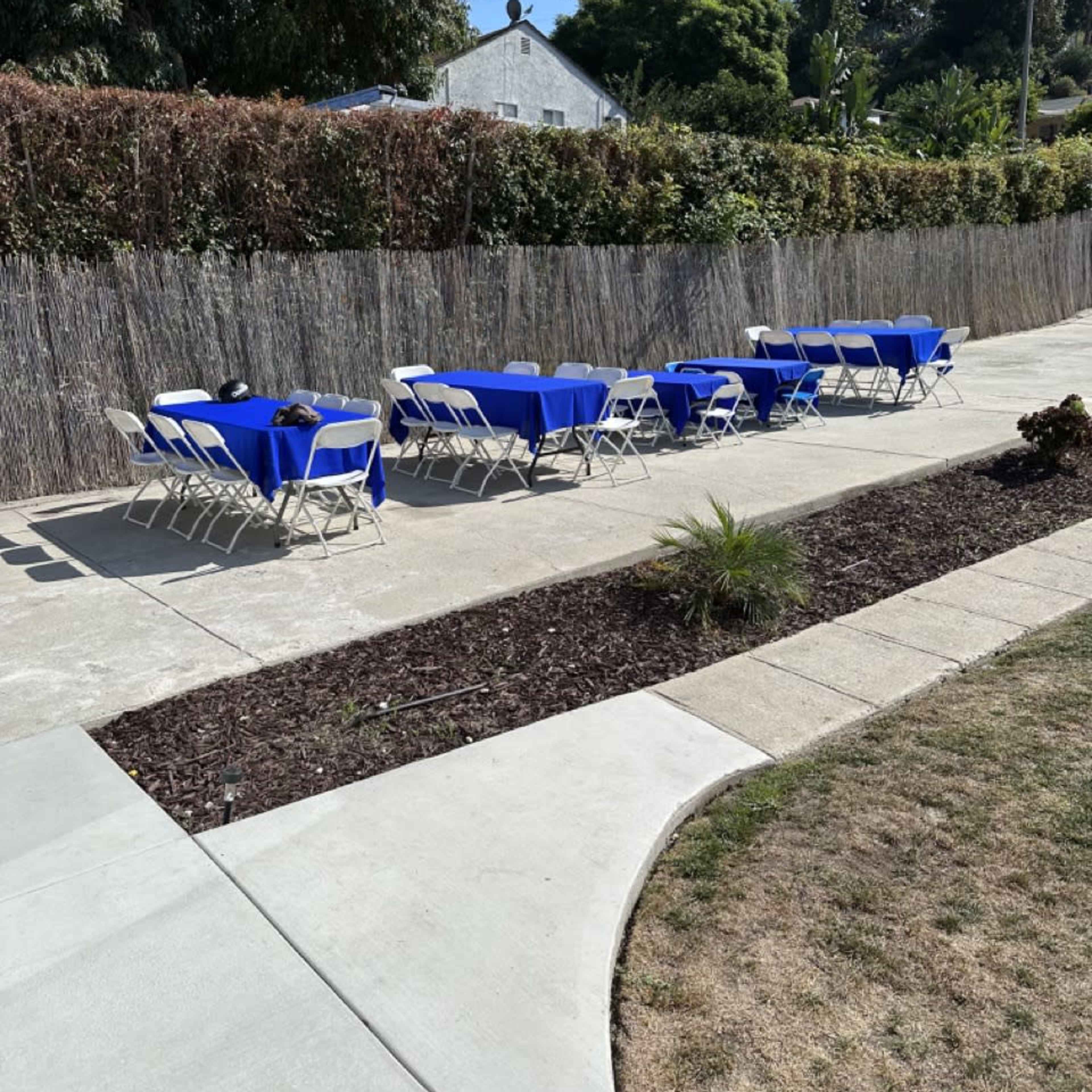 A patio area with several blue tables and chairs arranged in two rows along a concrete pathway, surrounded by greenery and a fence.