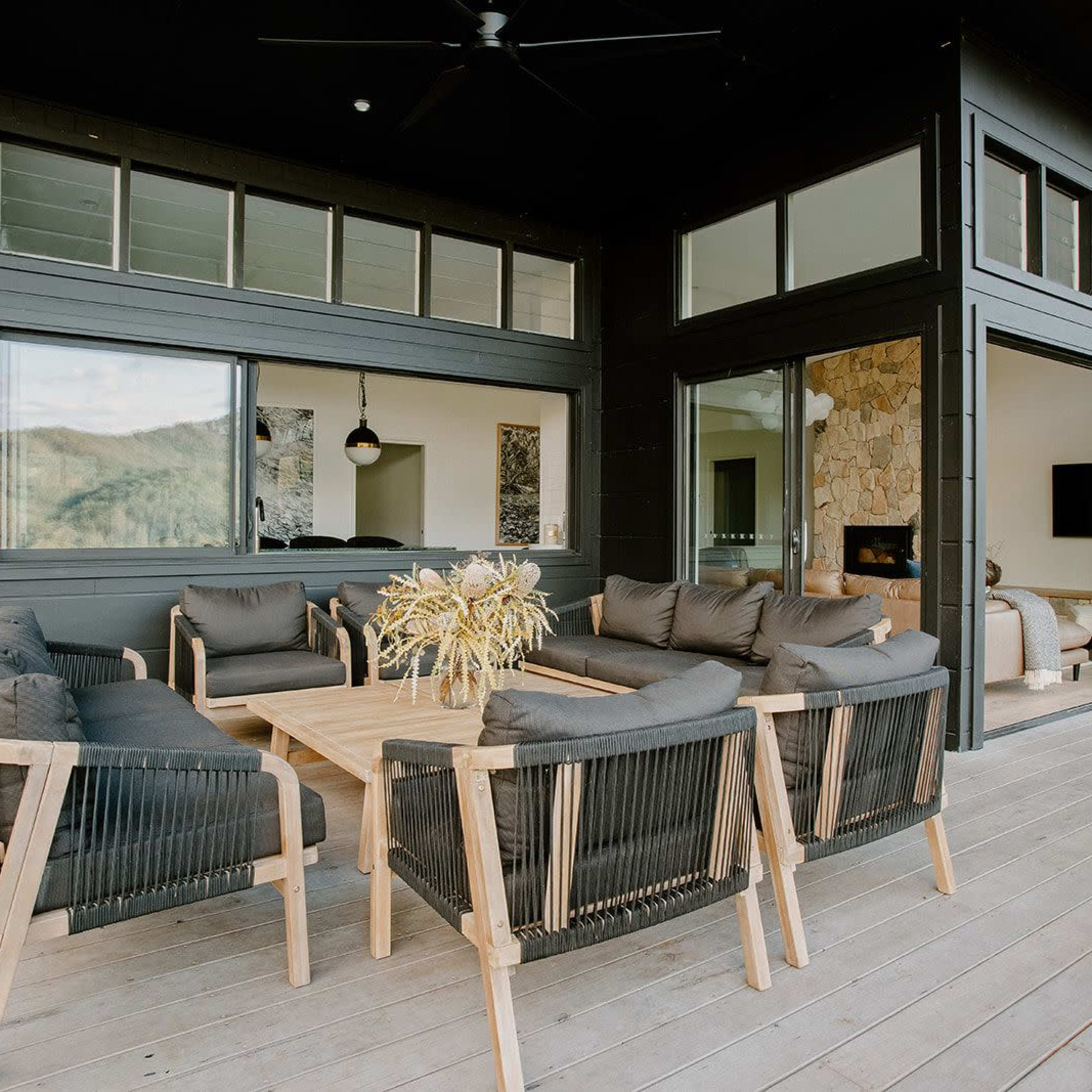 A modern outdoor seating area features a wooden table surrounded by black cushioned chairs against a backdrop of large glass windows.