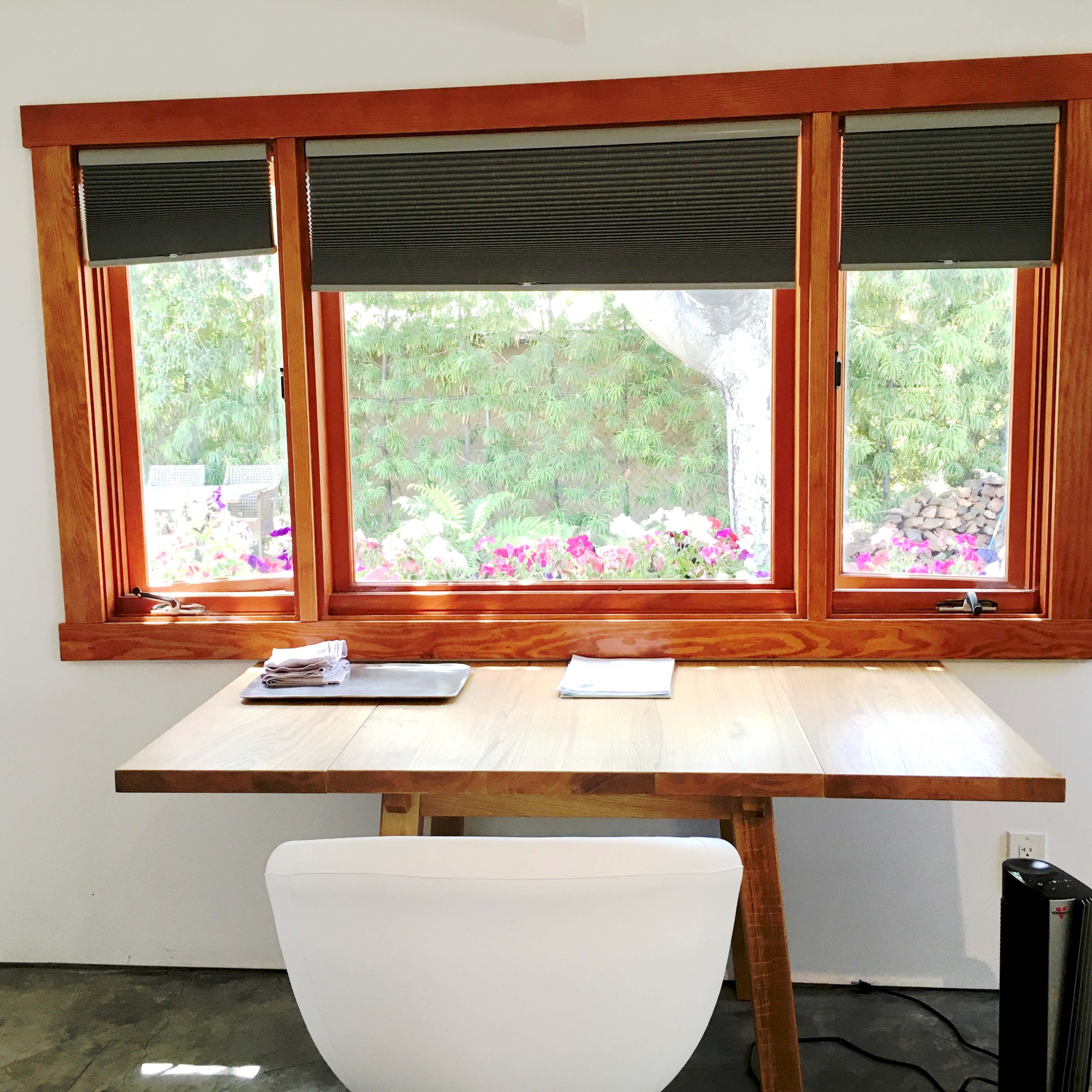 A wooden desk with a white chair is positioned near a window featuring wooden frames and roller shades, overlooking a garden with flowers.