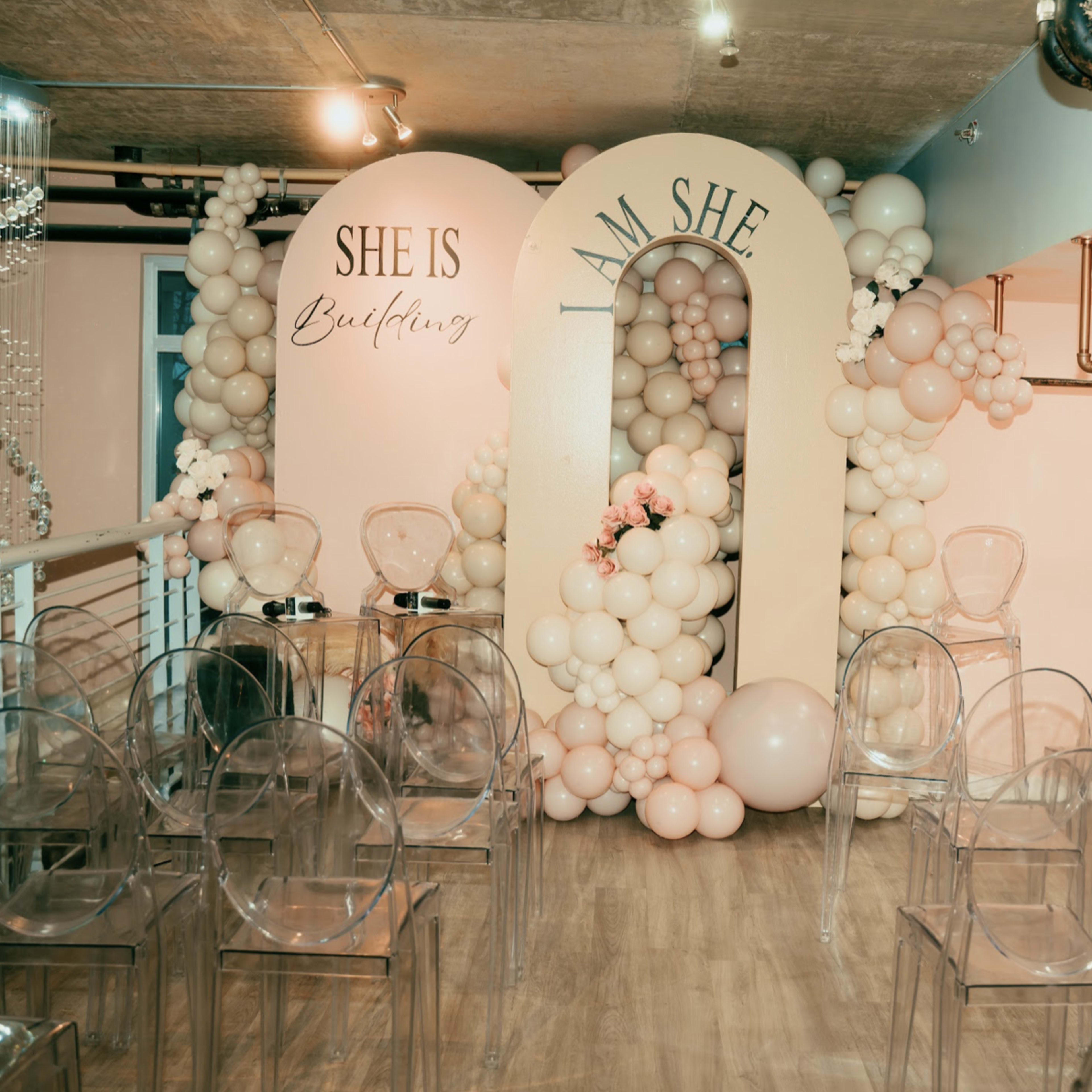 A decorated event space featuring an arch adorned with balloons, flanked by two signs labeled "SHE IS" and "I AM SHE," with rows of clear chairs arranged in front.
