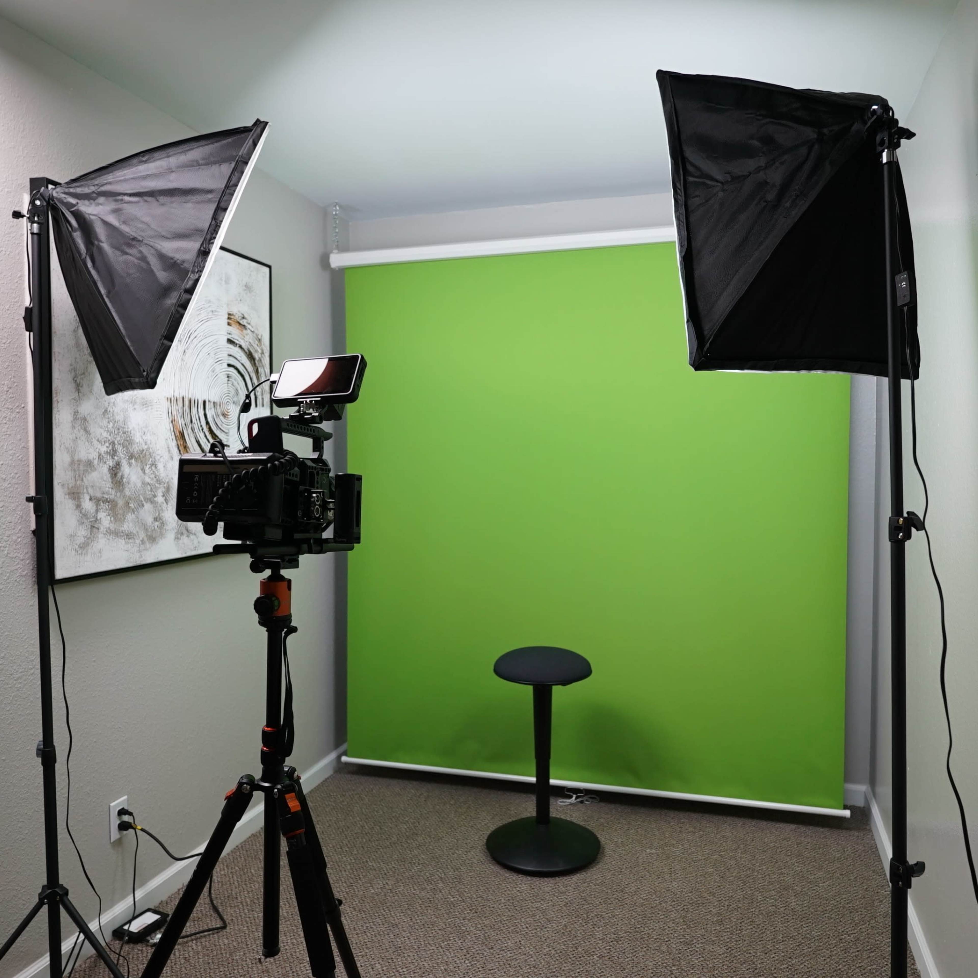 A small studio setup with a camera on a tripod, two softbox lights, a green backdrop, and a black stool.