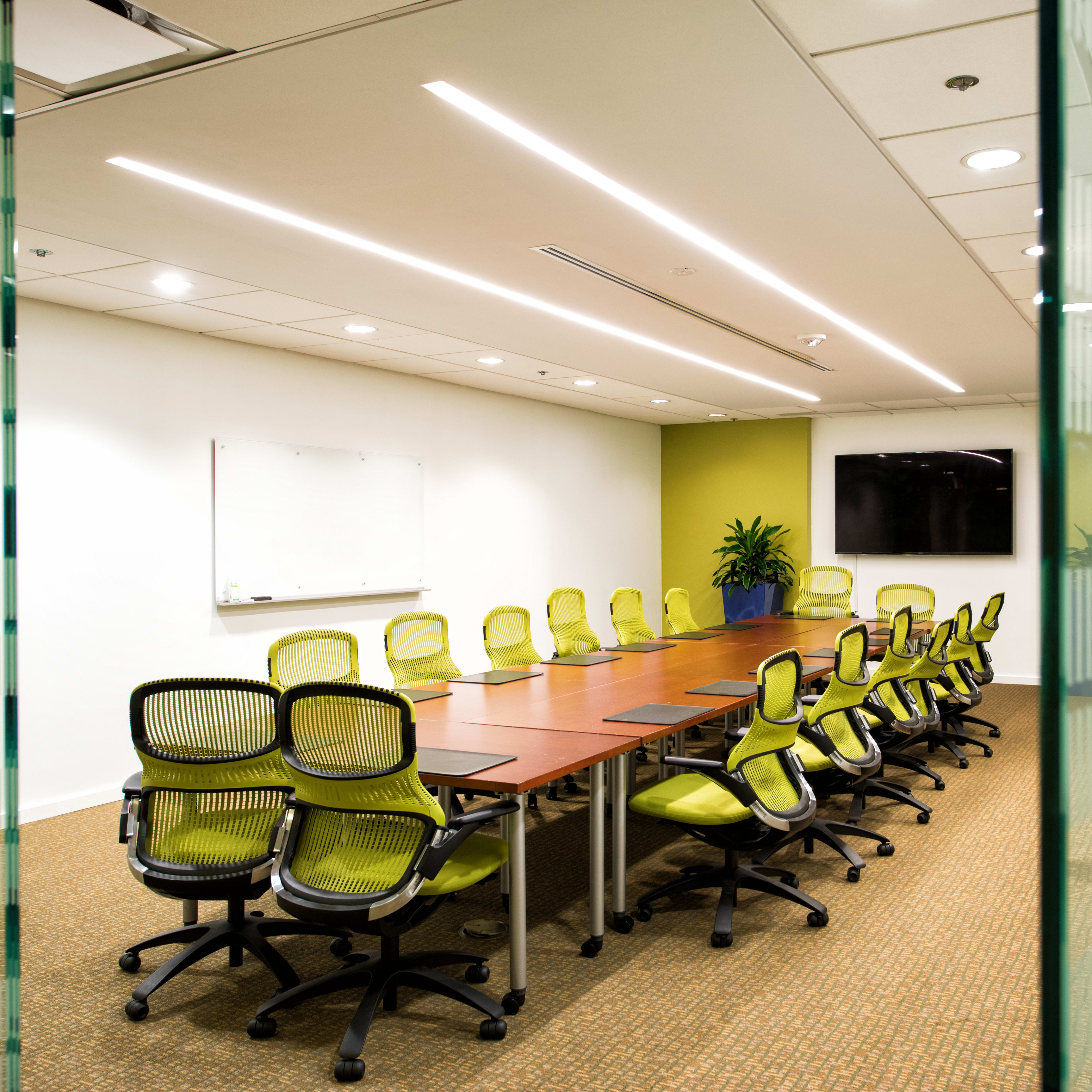 A large conference room features a long wooden table surrounded by green ergonomic chairs, with a whiteboard and a TV on the wall.