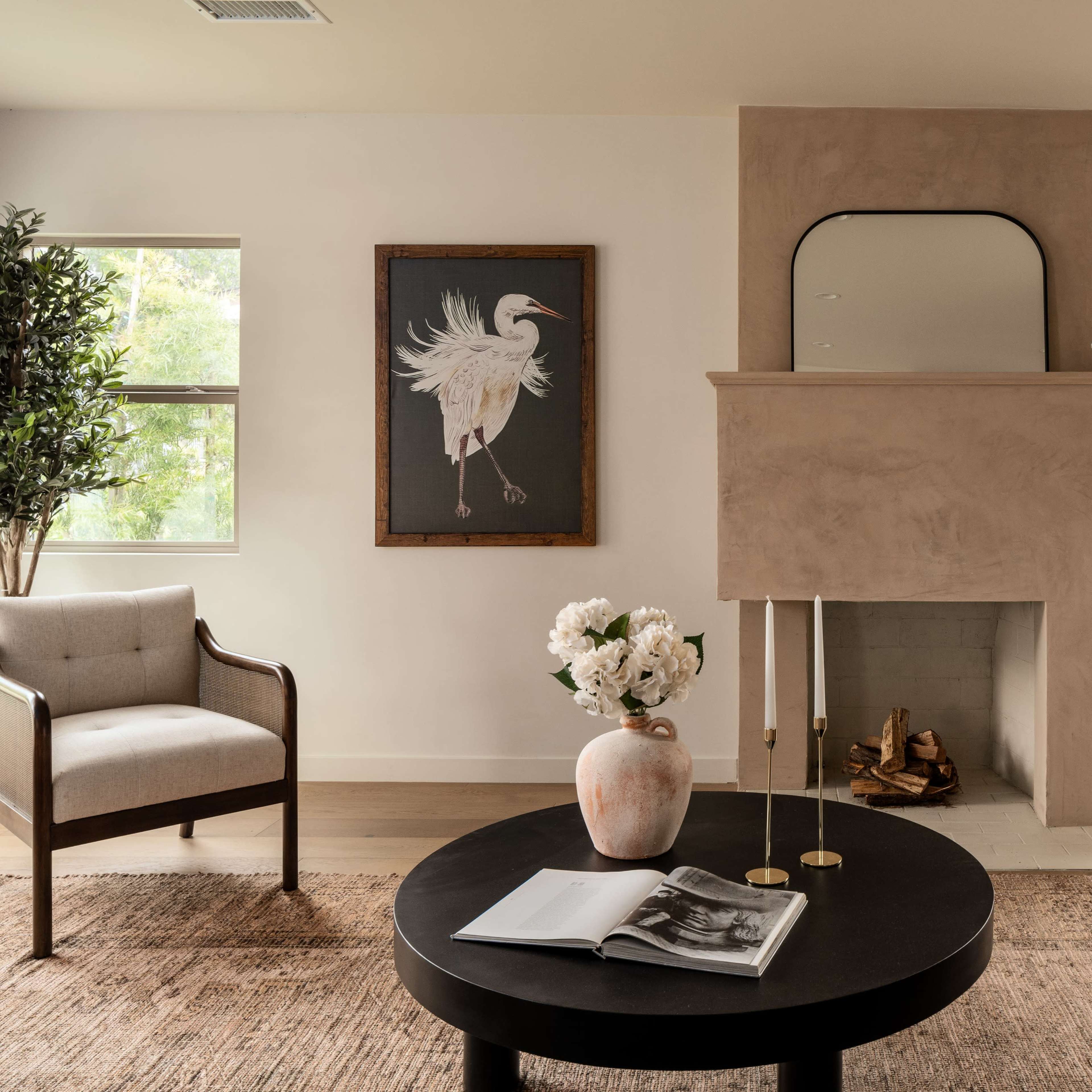 A modern living room with a minimalist design featuring a fireplace, a round coffee table with a book, and a framed artwork of a bird on the wall.