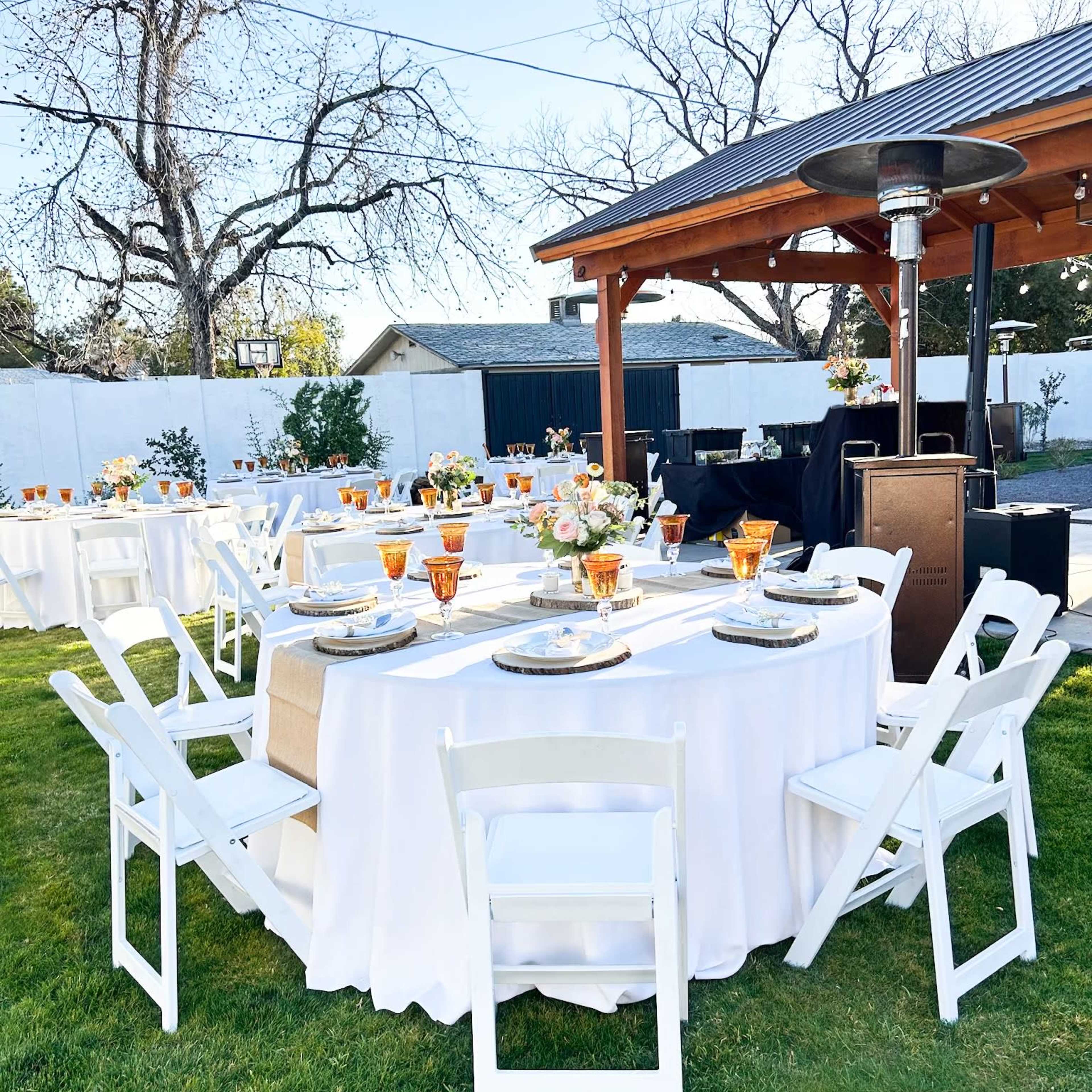 A set of elegantly arranged tables with white tablecloths and chairs in a green outdoor space, under a wooden gazebo with a solar panel.