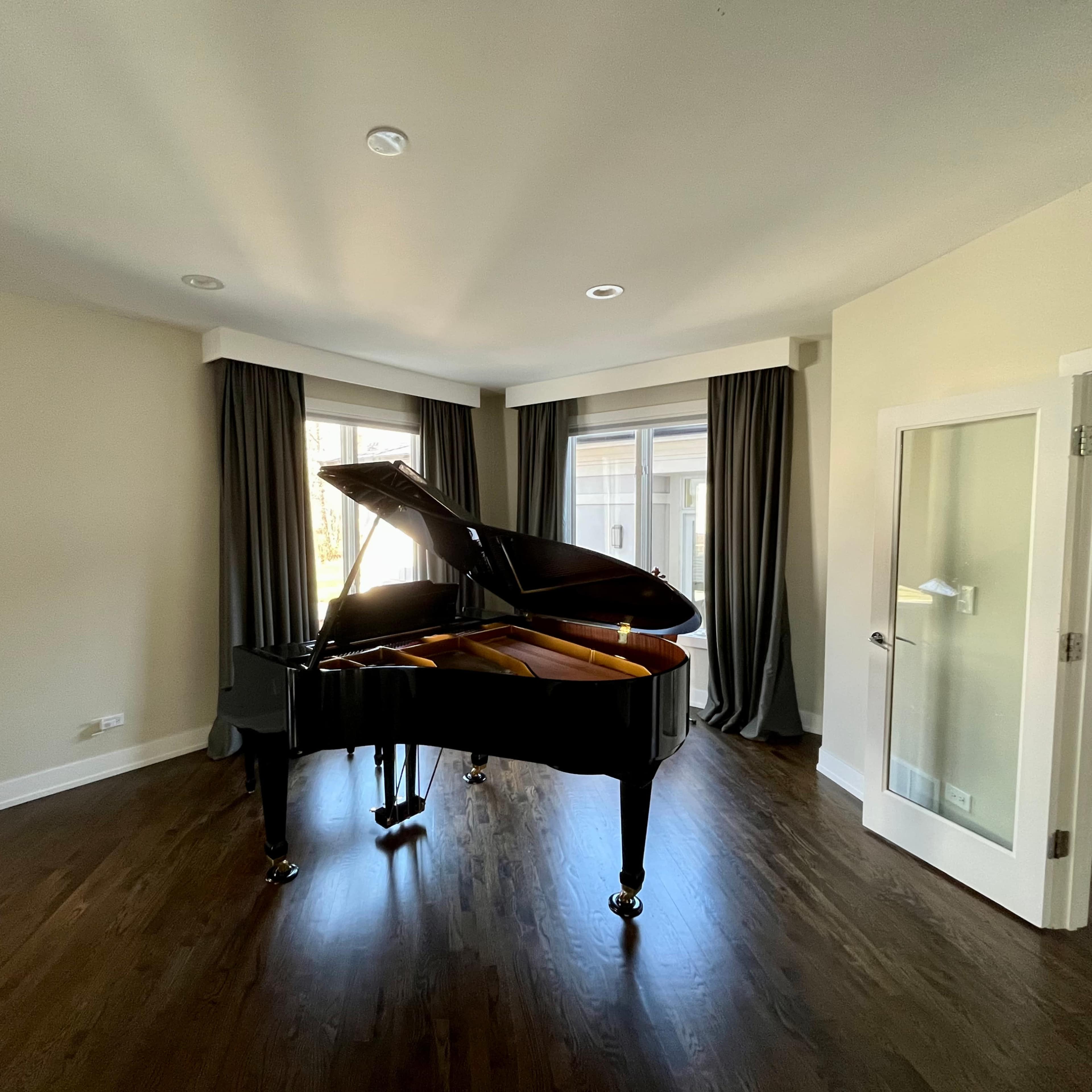 A grand piano is positioned in the center of a well-lit room with large windows and dark curtains.