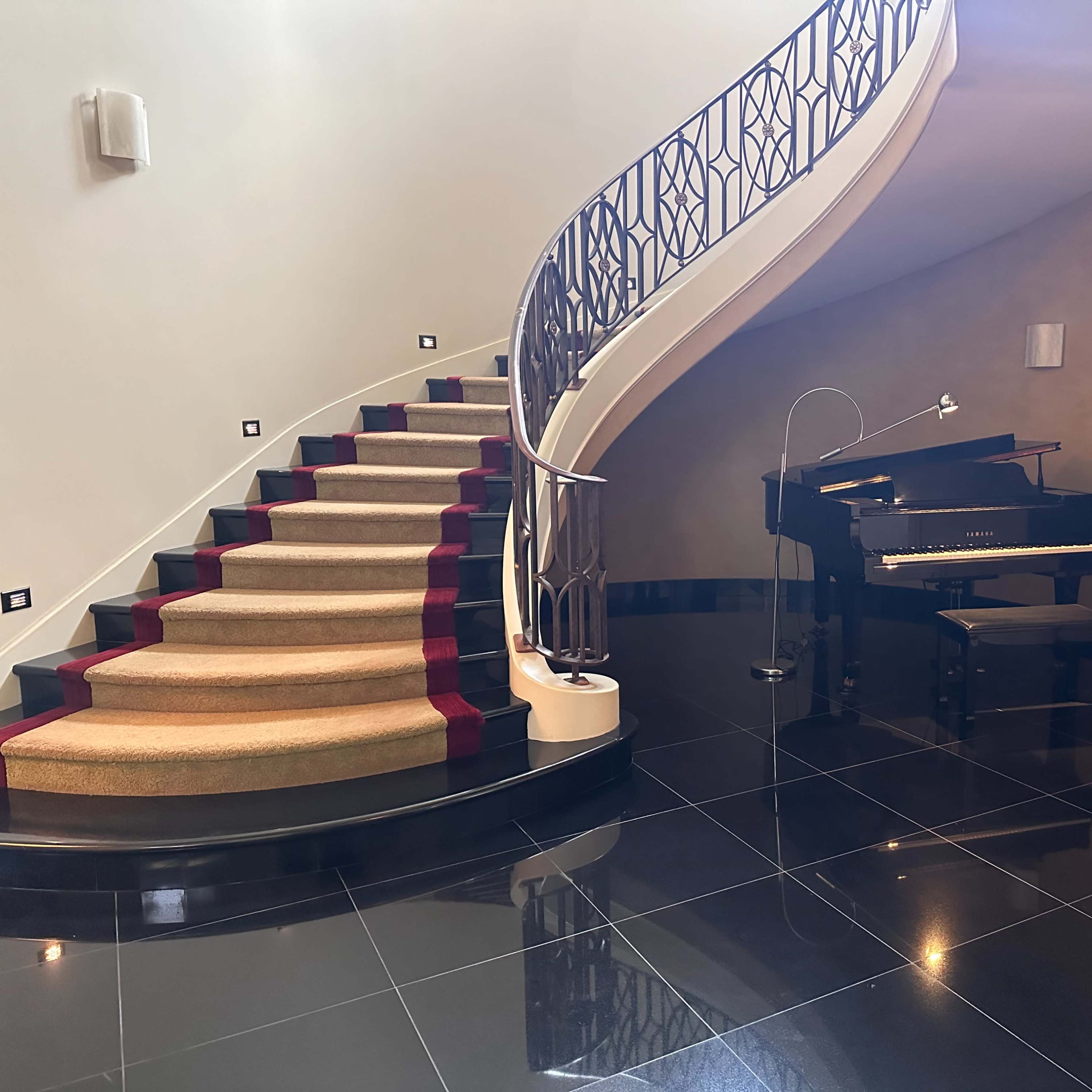 A spiral staircase leading to an upper level beside a black grand piano positioned on glossy tile flooring, with a painting of a flower on the wall.