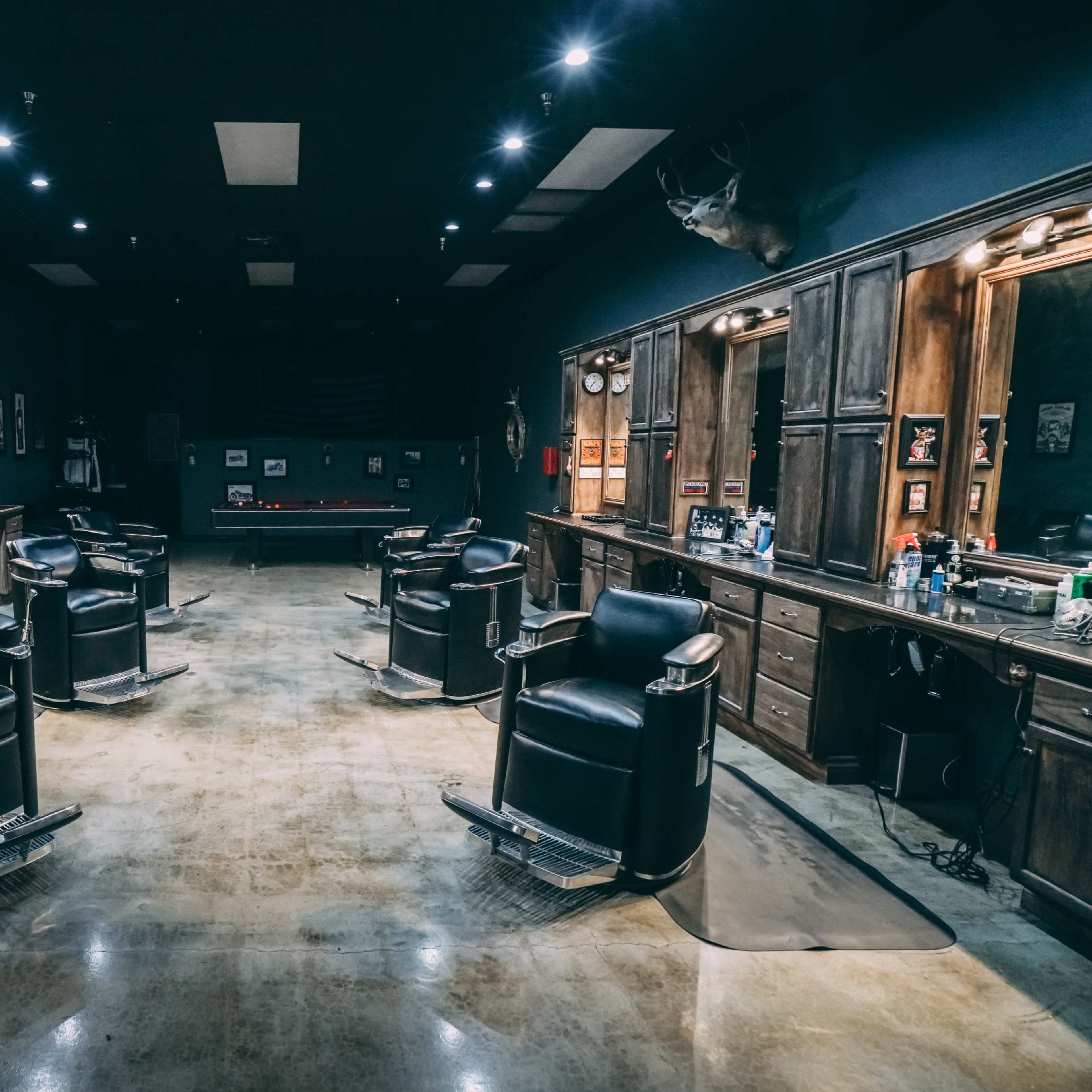 A barber shop with four black barber chairs arranged in a spacious, dimly lit room featuring wooden cabinetry and a pool table in the background.