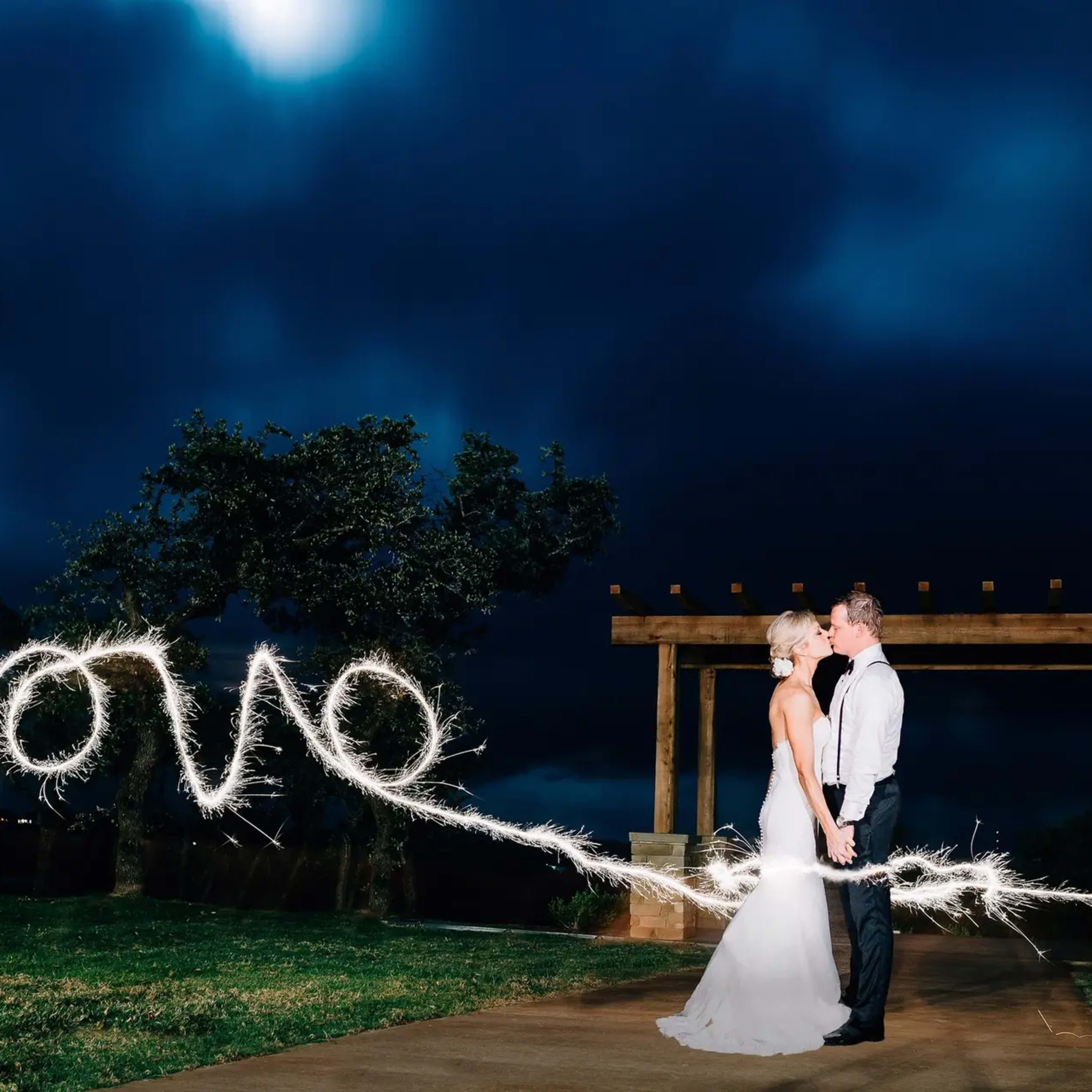A bride and groom stand together under a dark sky while holding sparklers that create the word "love."