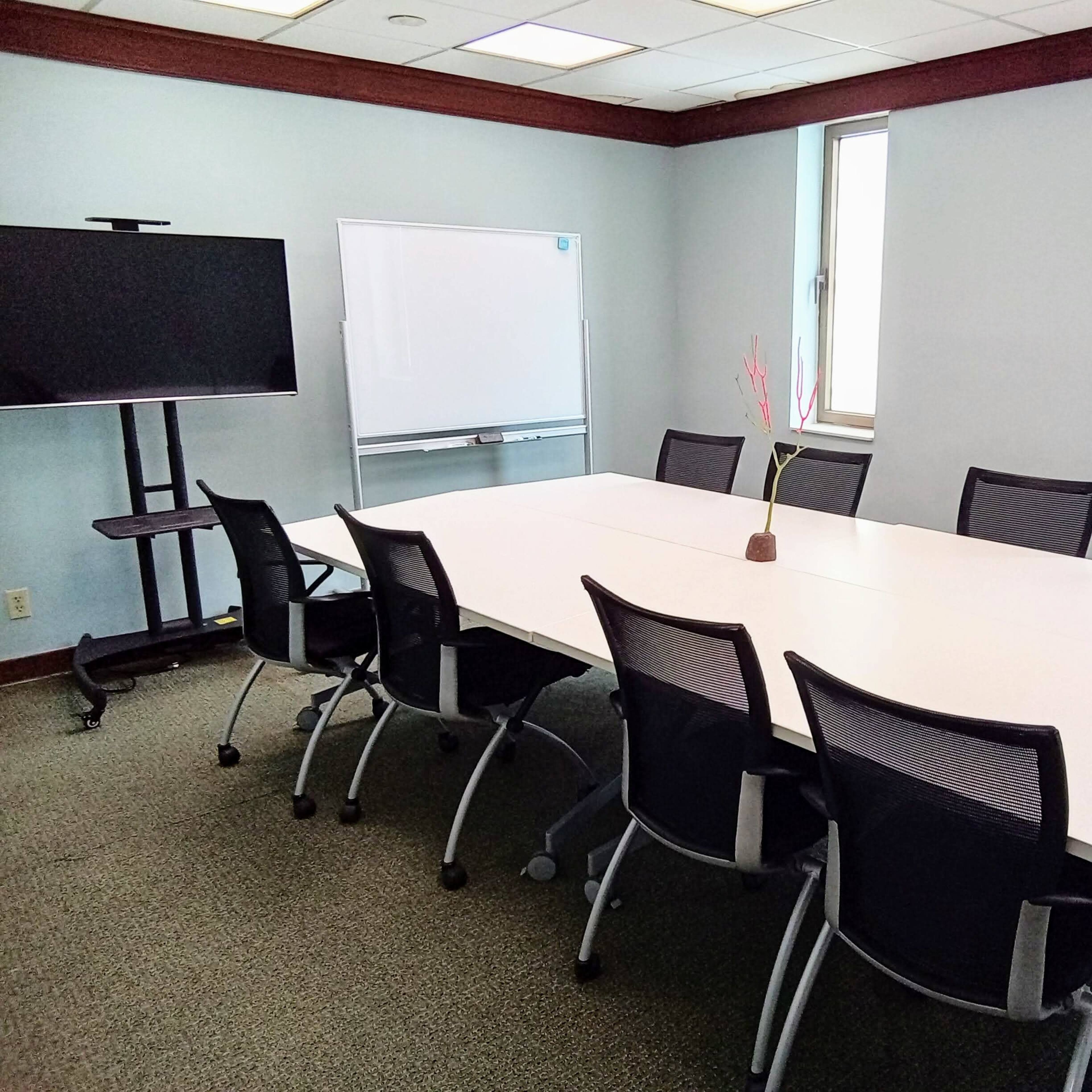 A modern conference room features a large rectangular table surrounded by eight chairs, a television on a stand, and a whiteboard against the wall.