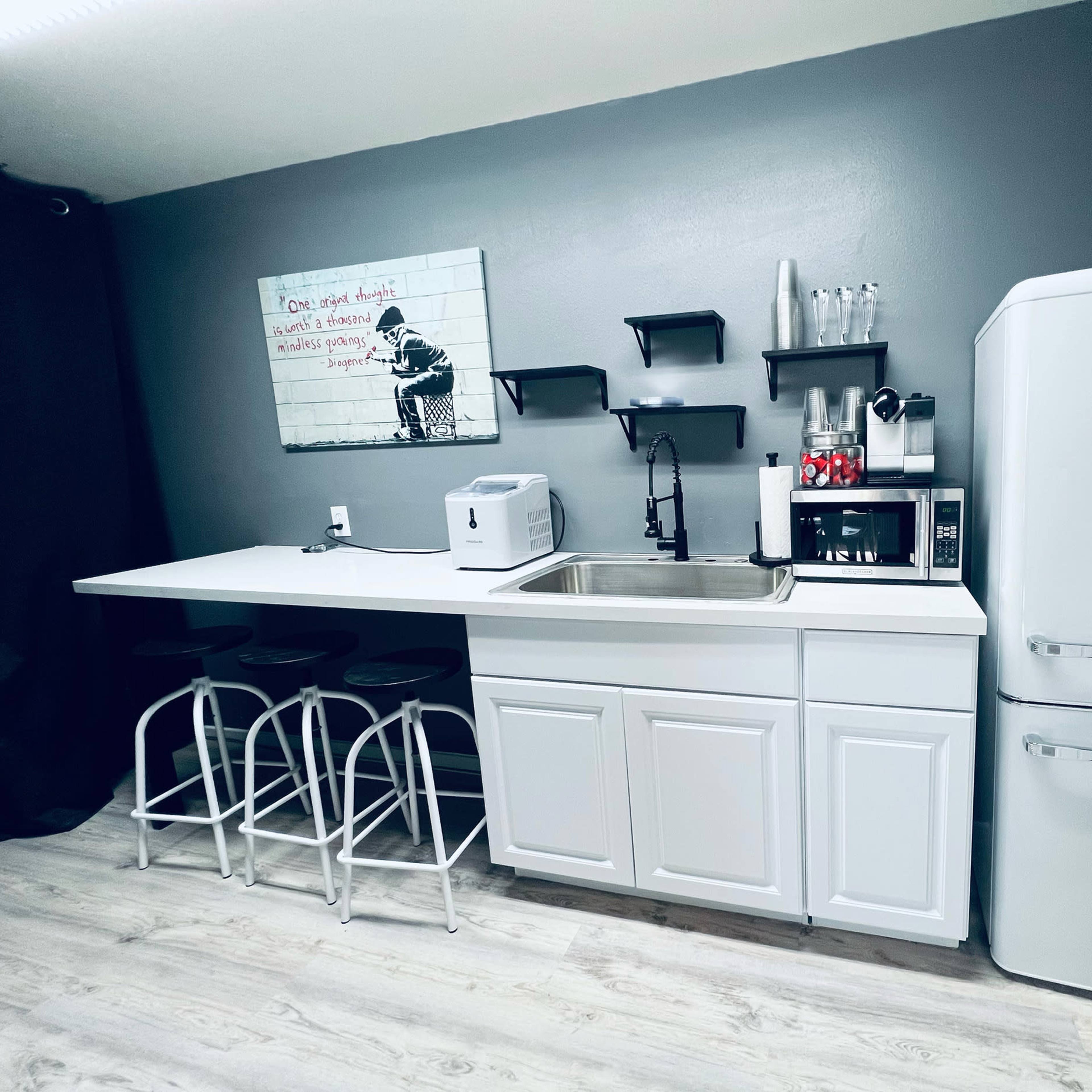 A modern kitchenette with white cabinetry, a stainless steel sink, a refrigerator, a microwave, and a coffee maker, set against a gray wall.