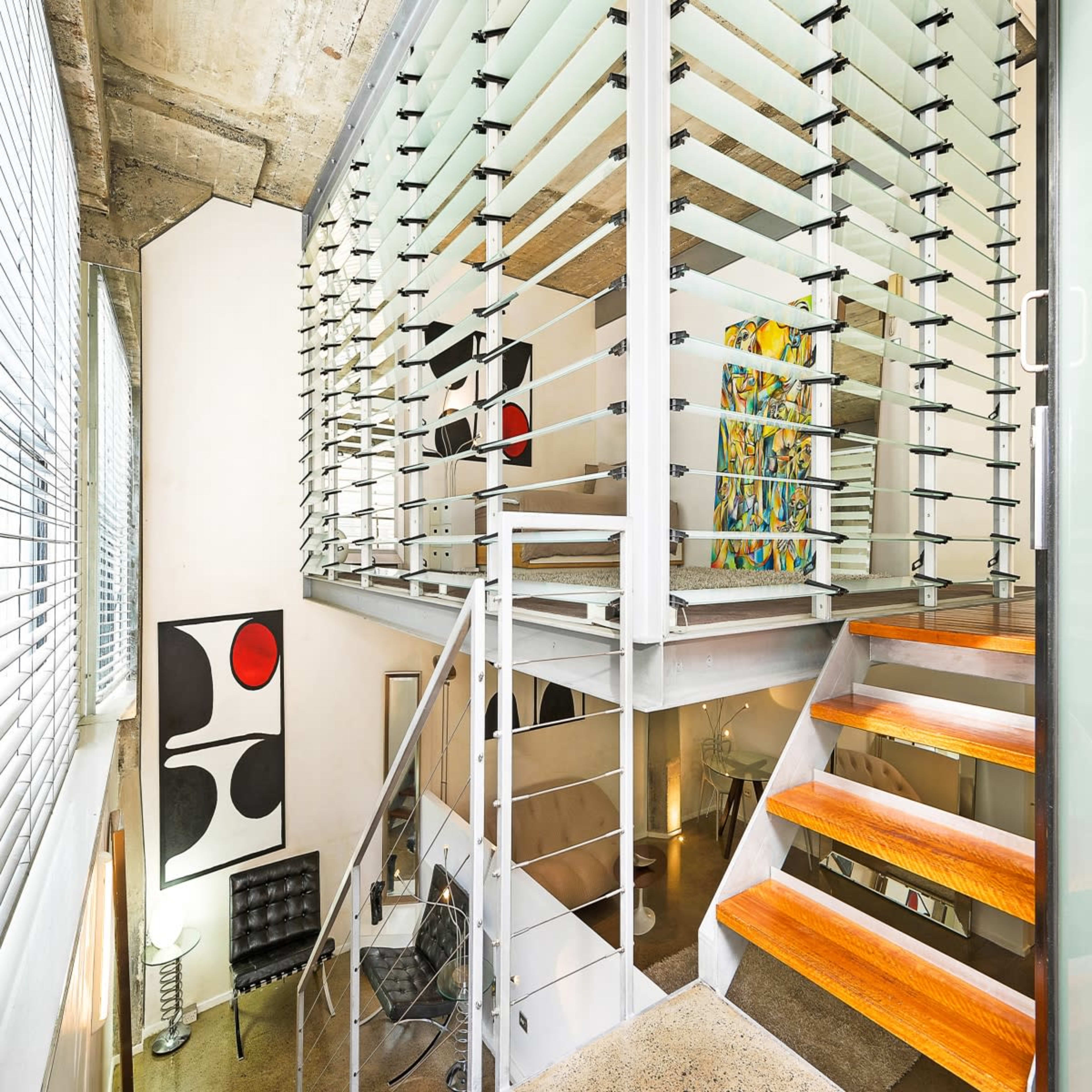 A modern, multi-level living space featuring glass walls, an open staircase with wooden steps, and contemporary art displayed on the walls.