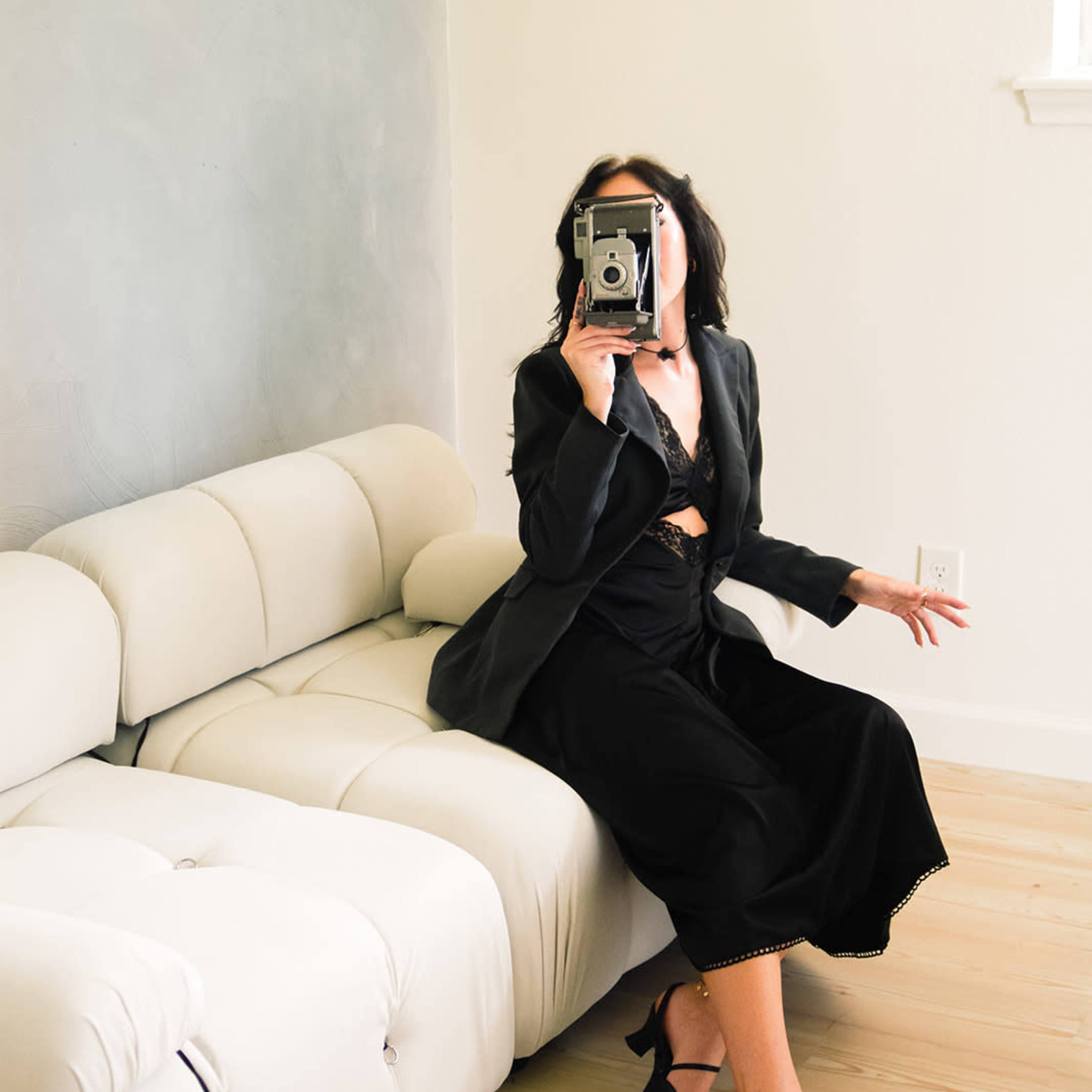 A woman in a black outfit sits on a light-colored sofa, holding a vintage camera towards the viewer.