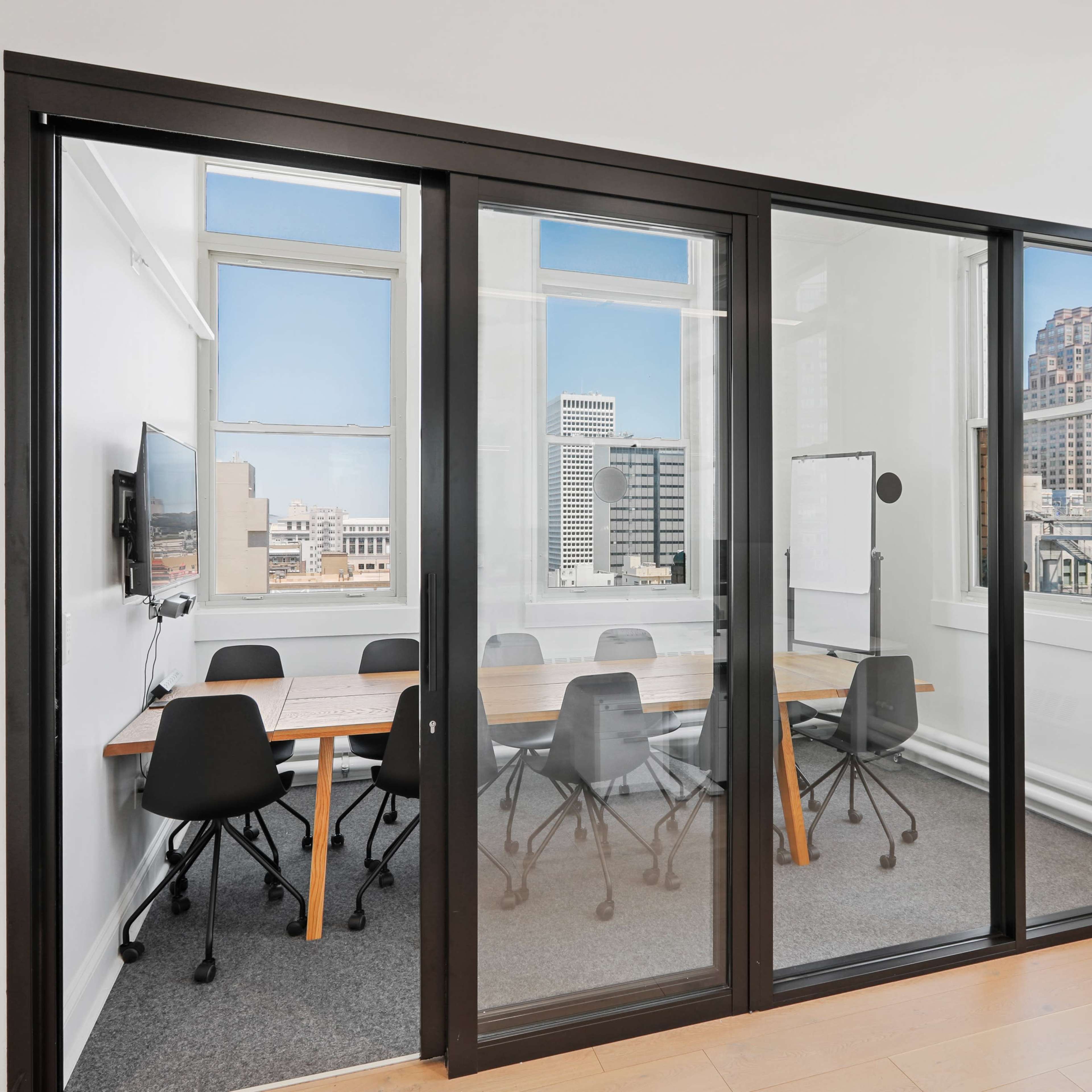 A modern glass-walled conference room features a large table surrounded by ergonomic chairs and offers a view of a cityscape through its windows.