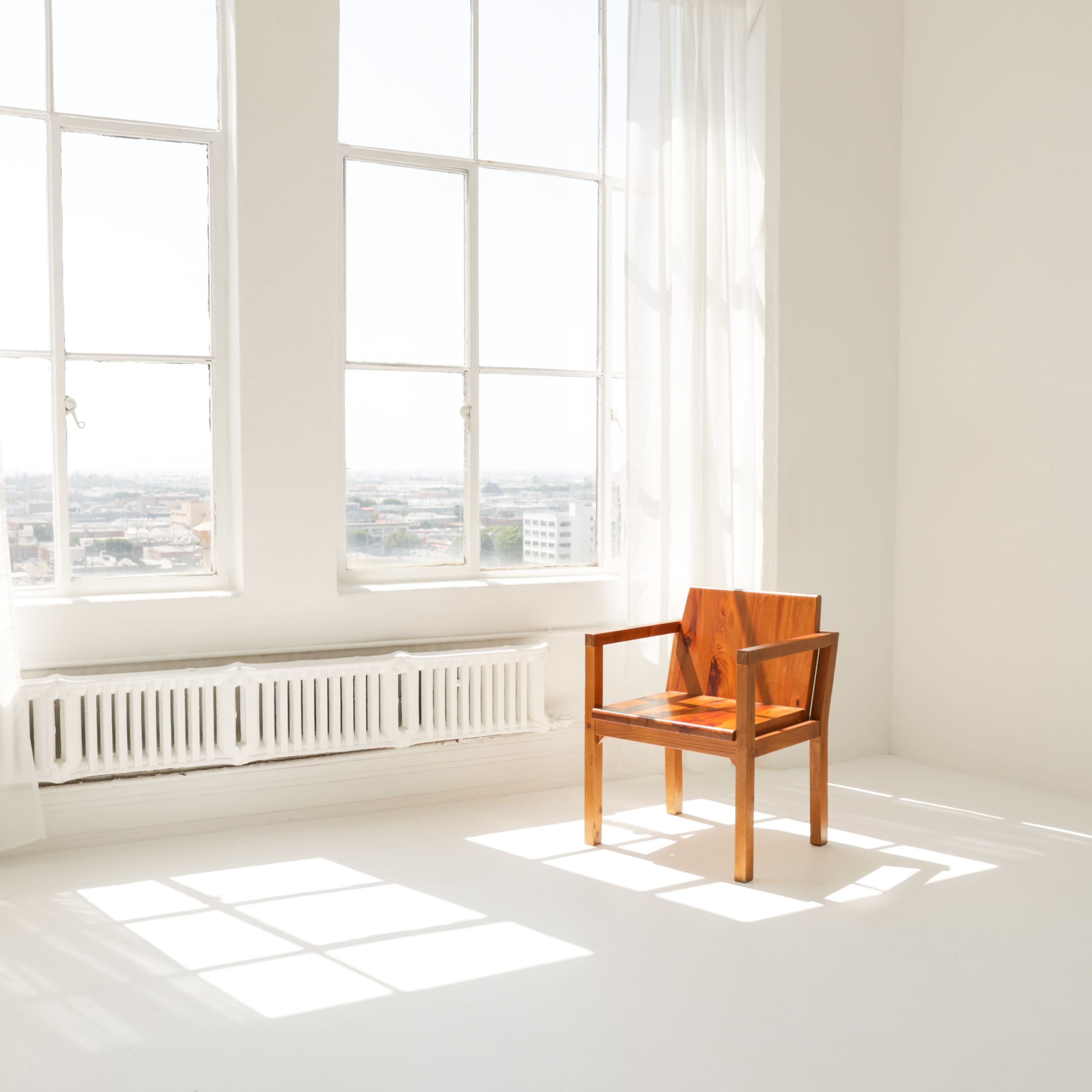 A wooden chair is placed in a bright, empty room with large windows that let in sunlight.