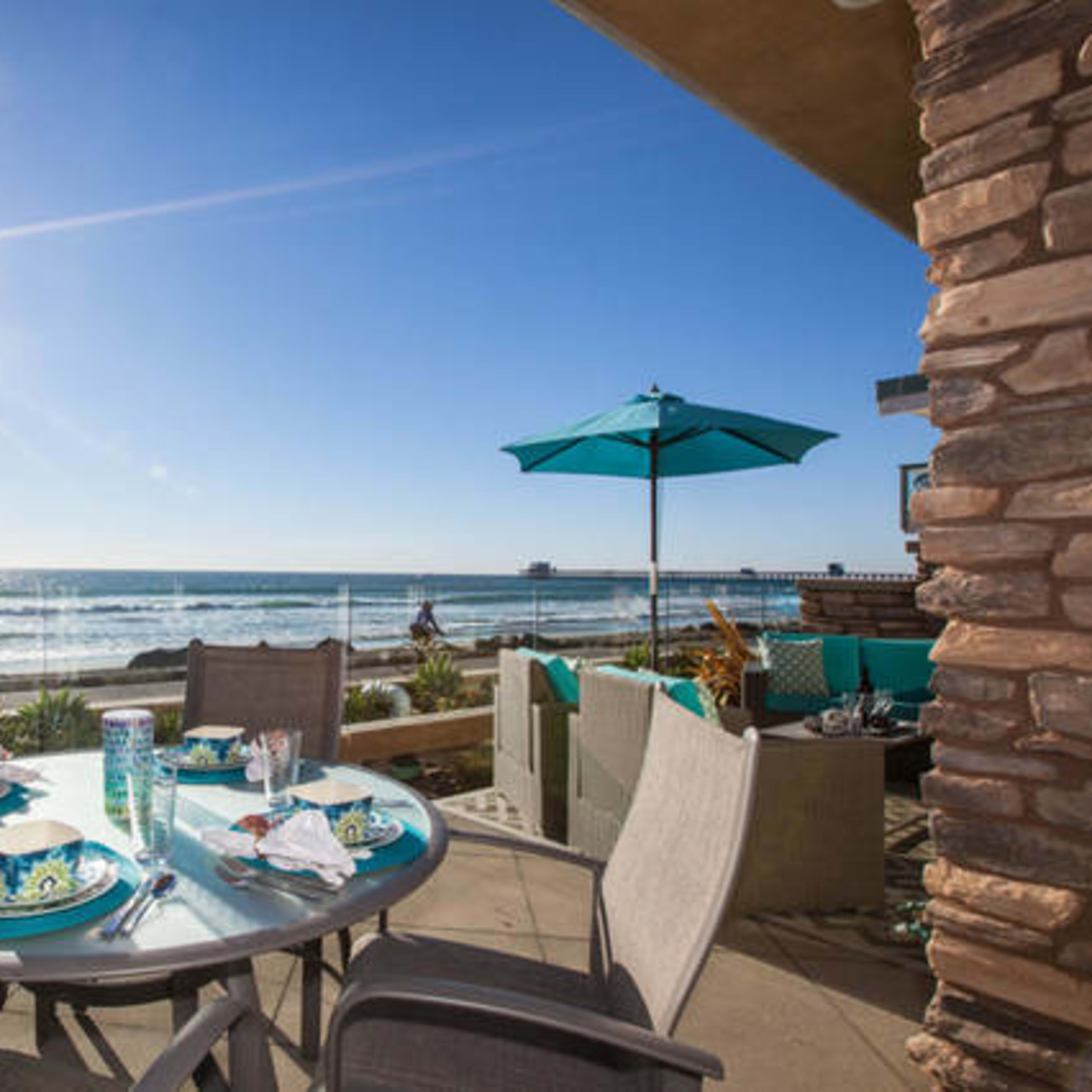 A patio dining set is positioned near the beach, overlooking the ocean with sun shining brightly in the background.