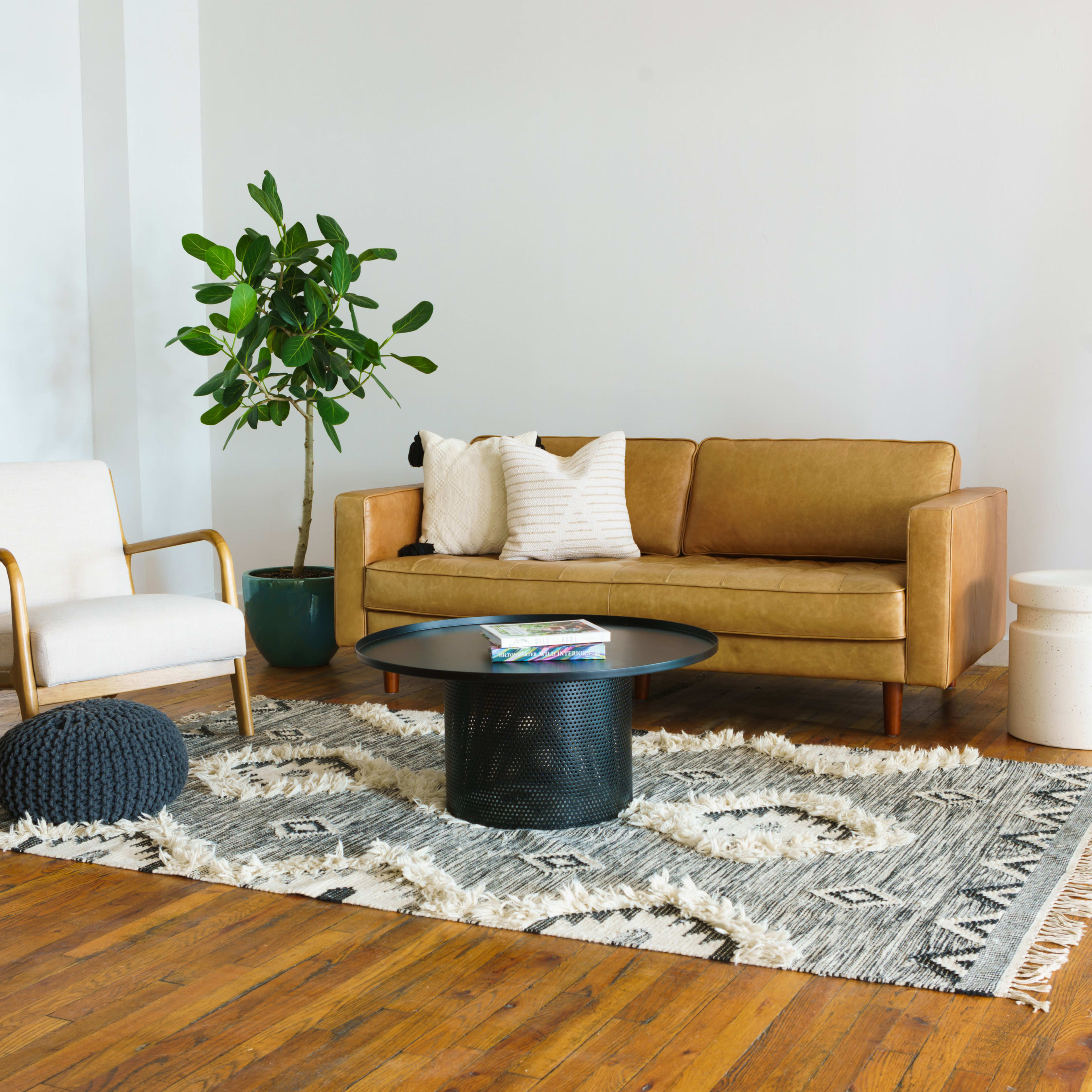 A modern living room features a tan leather sofa, a white armchair, a round black coffee table, a patterned rug, and various potted plants.