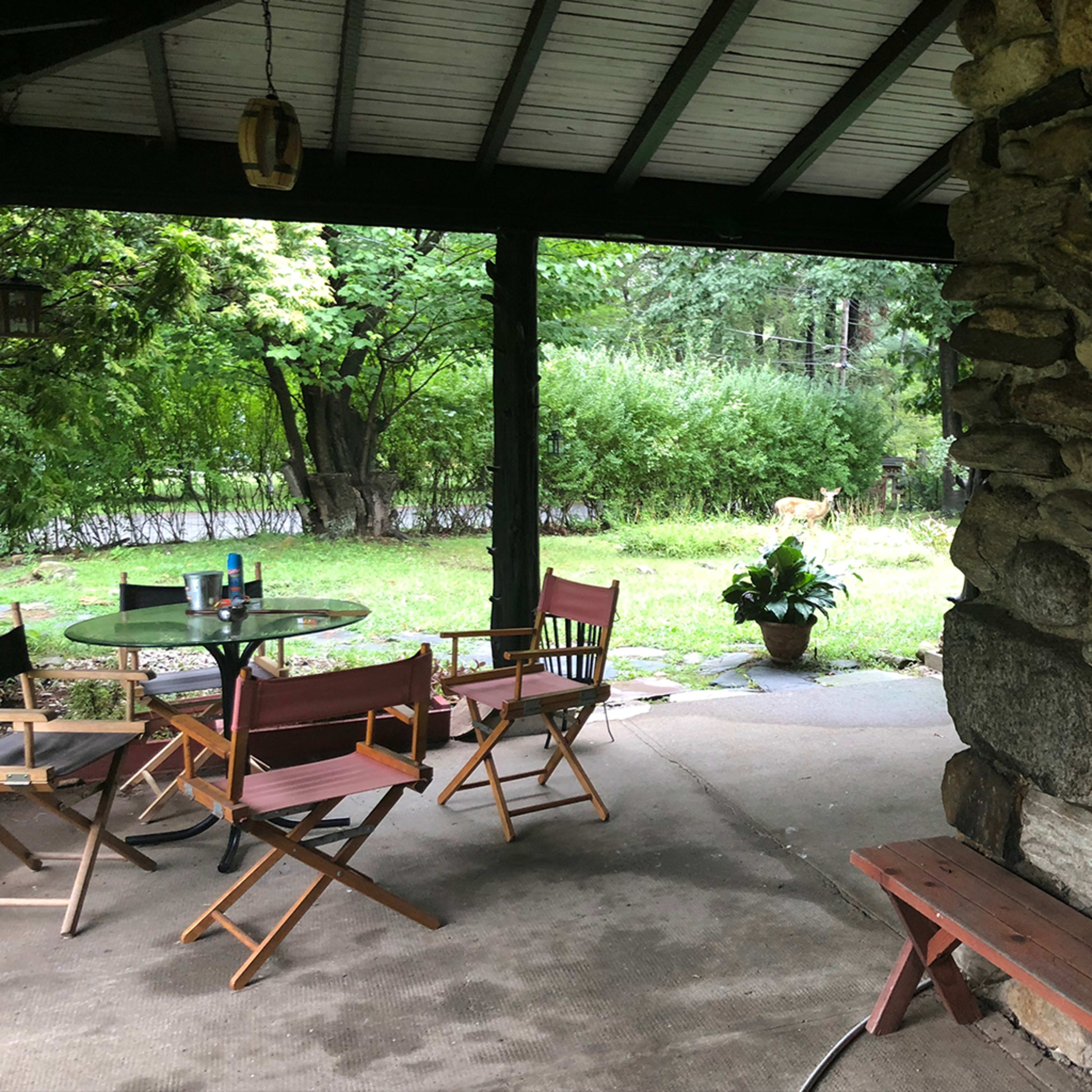 A stone wall supports a covered patio with wooden chairs arranged around a glass table, overlooking a grassy area with trees in the background.