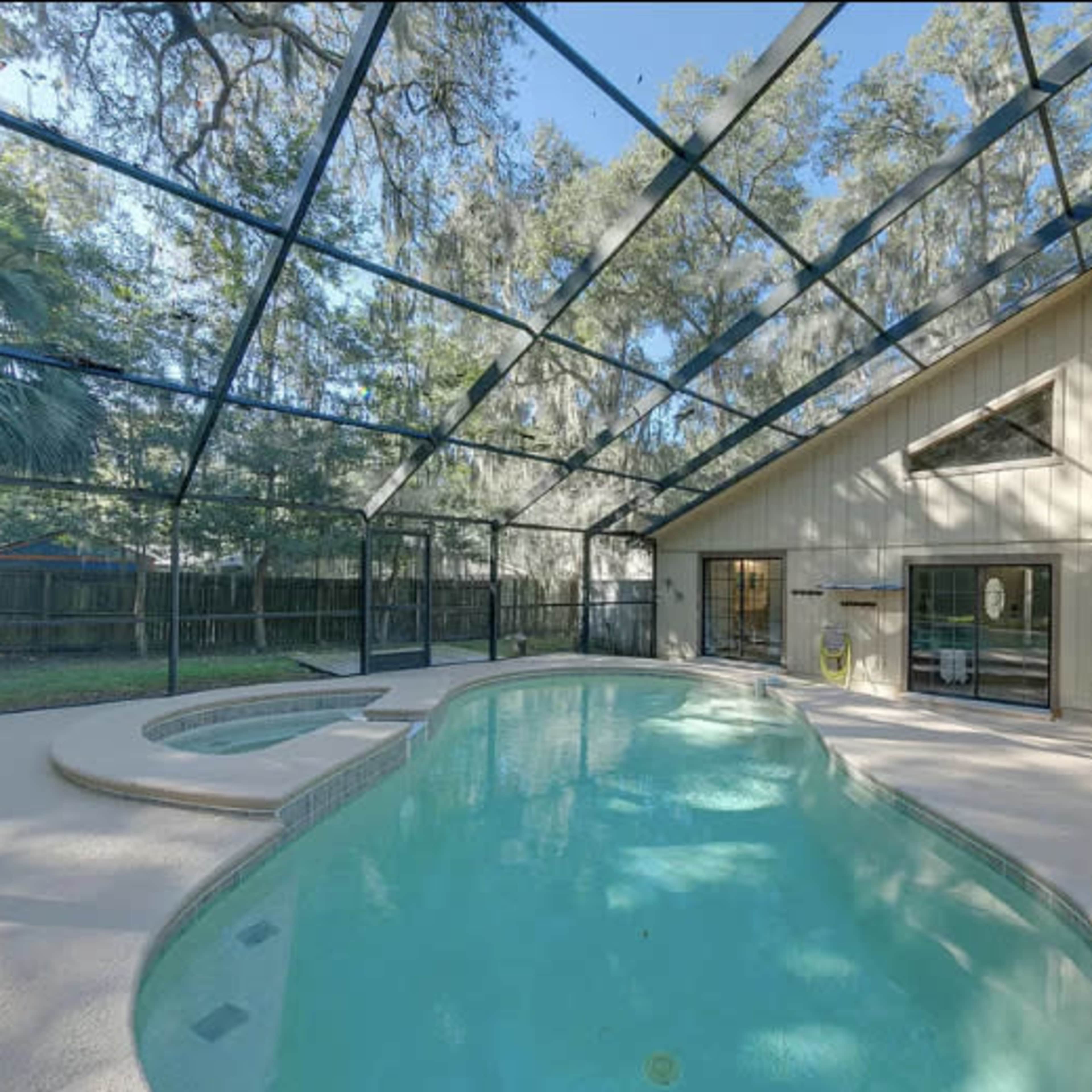 A screened-in pool area featuring a swimming pool with a spa, surrounded by greenery and a home with large glass doors.