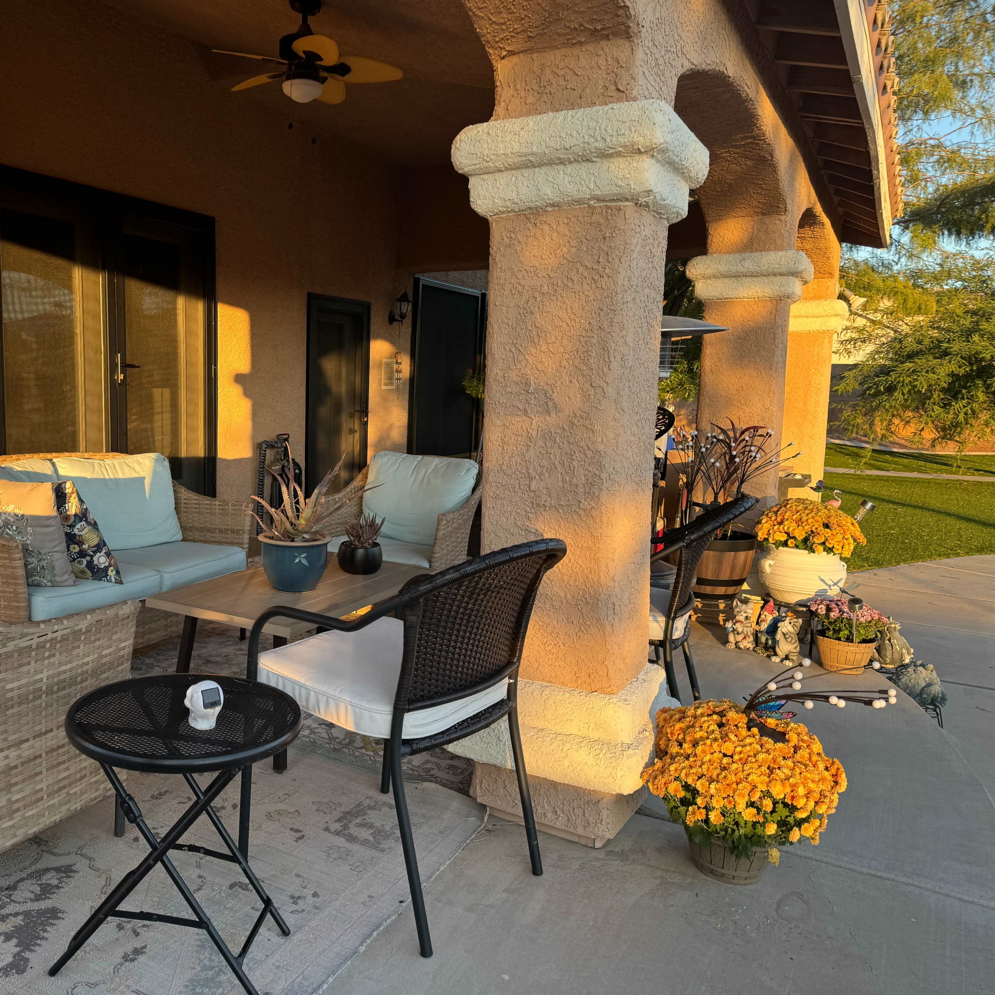 A cozy outdoor seating area features a wicker sofa, chairs, and a small table, surrounded by potted flowers and illuminated by warm afternoon light.