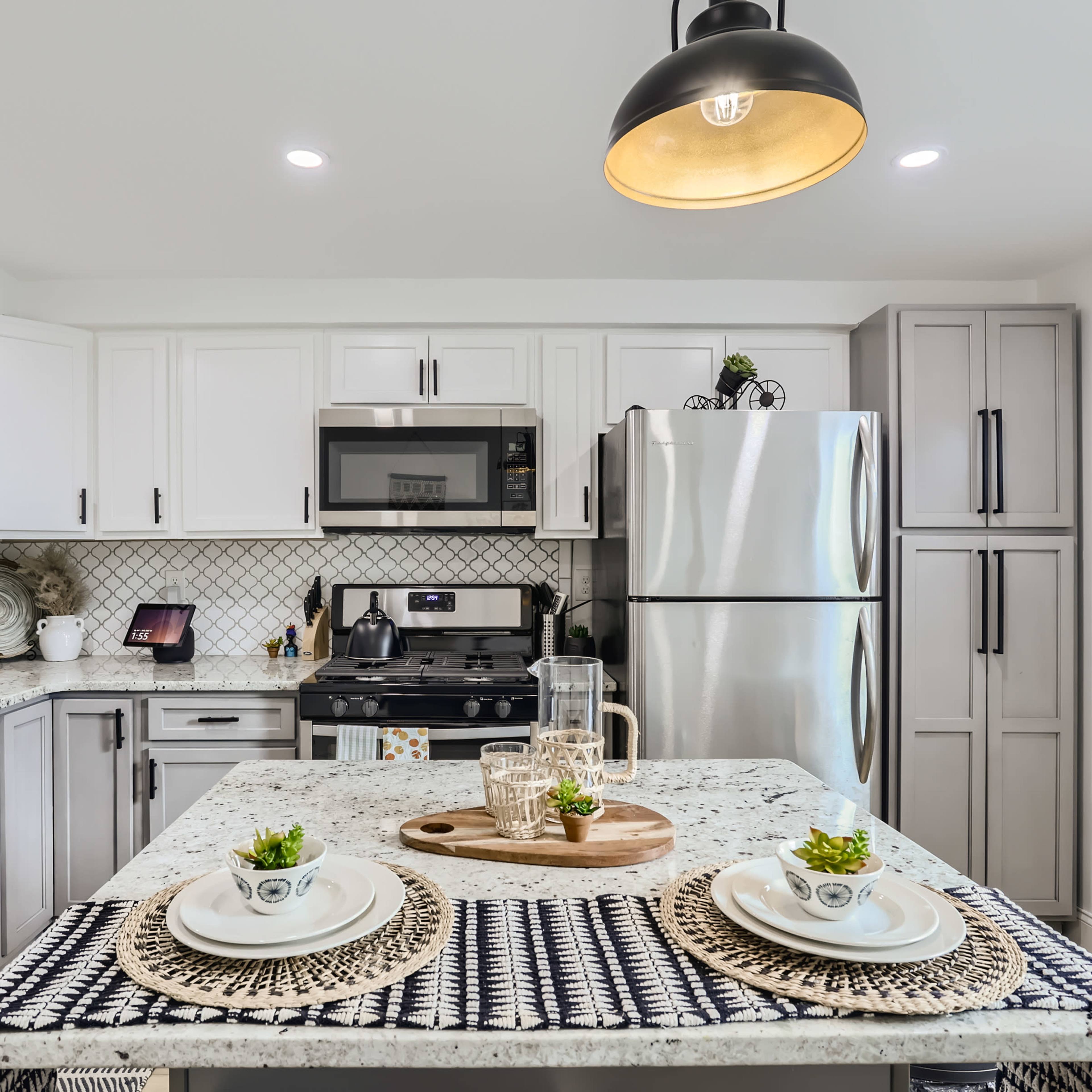 A modern kitchen features a central island with two place settings, stainless steel appliances, and a combination of light and dark cabinetry.
