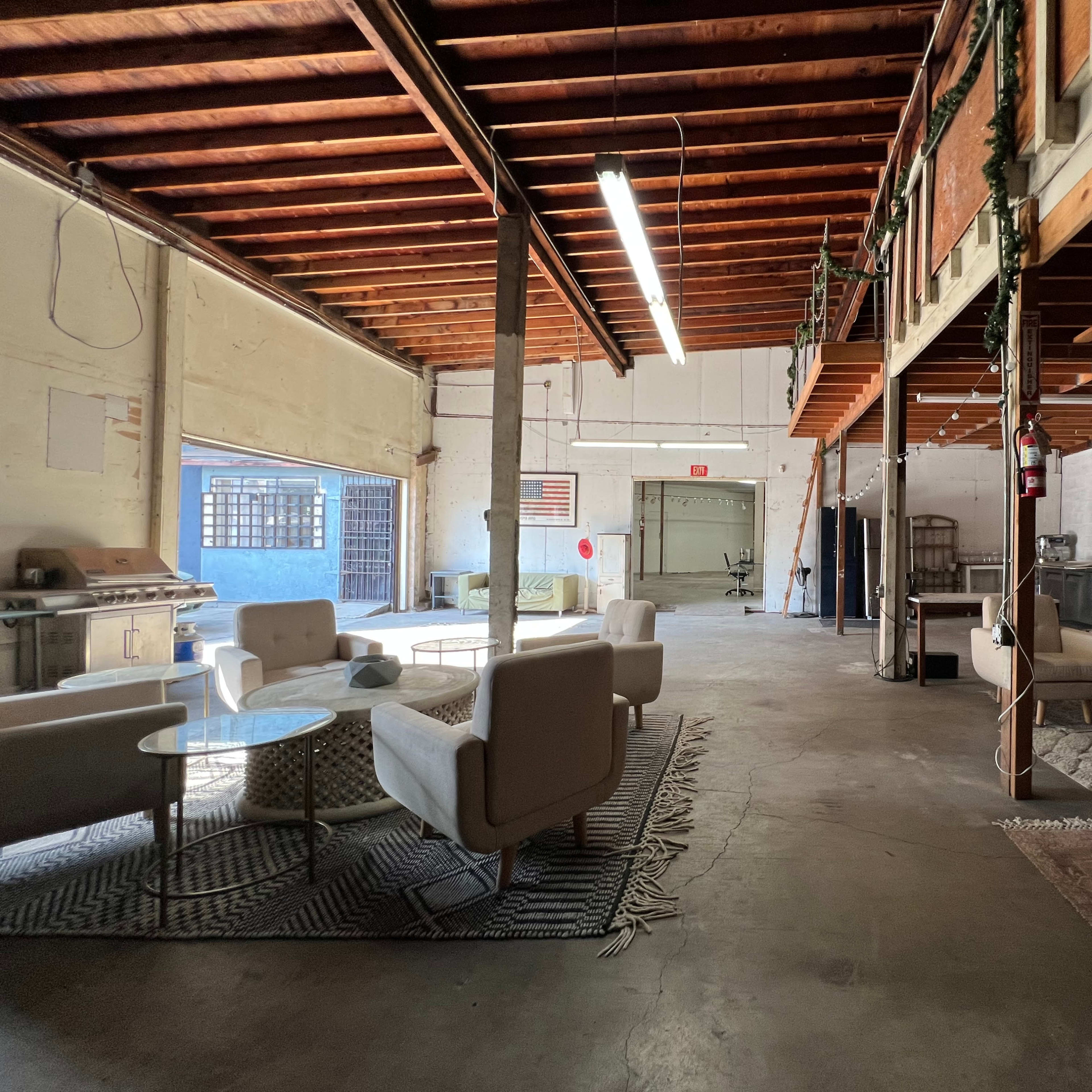 An open warehouse space with a seating area featuring modern chairs and tables, illuminated by overhead lights.