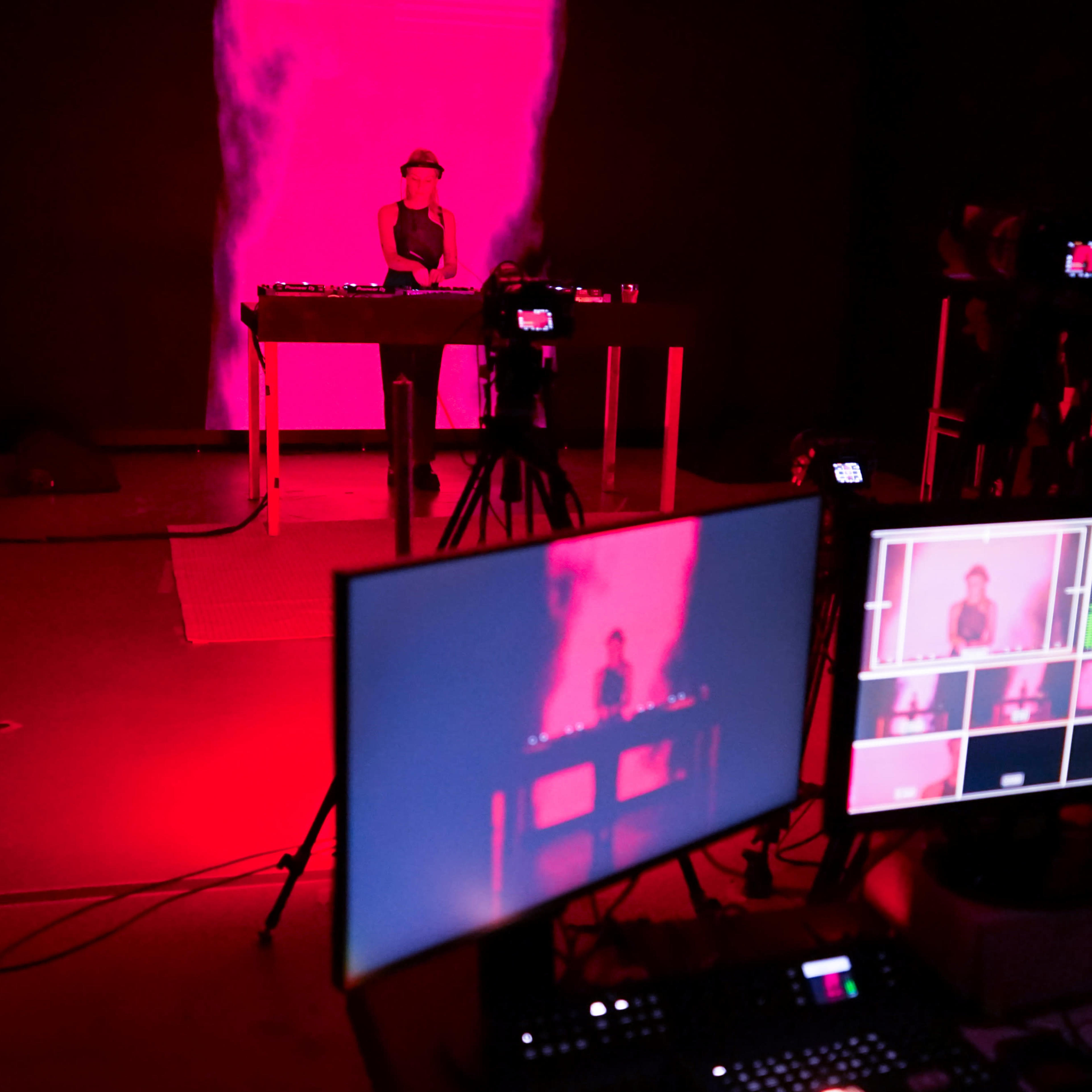 A person stands behind a DJ set in a dimly lit room illuminated by red lights, with cameras and monitors set up to capture the performance.