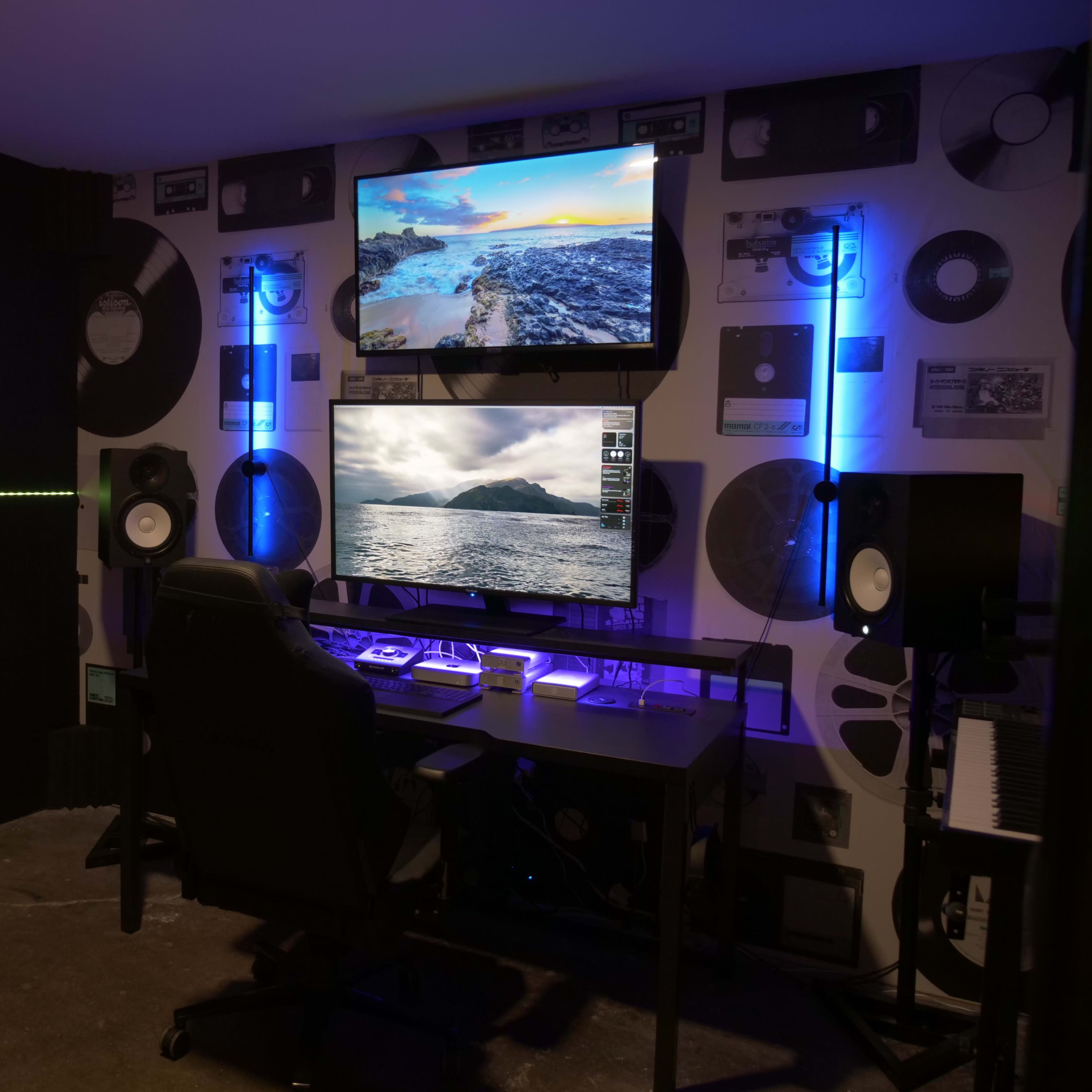A modern workspace features a large desk with dual monitors displaying landscapes, surrounded by vinyl records on the wall and ambient lighting.