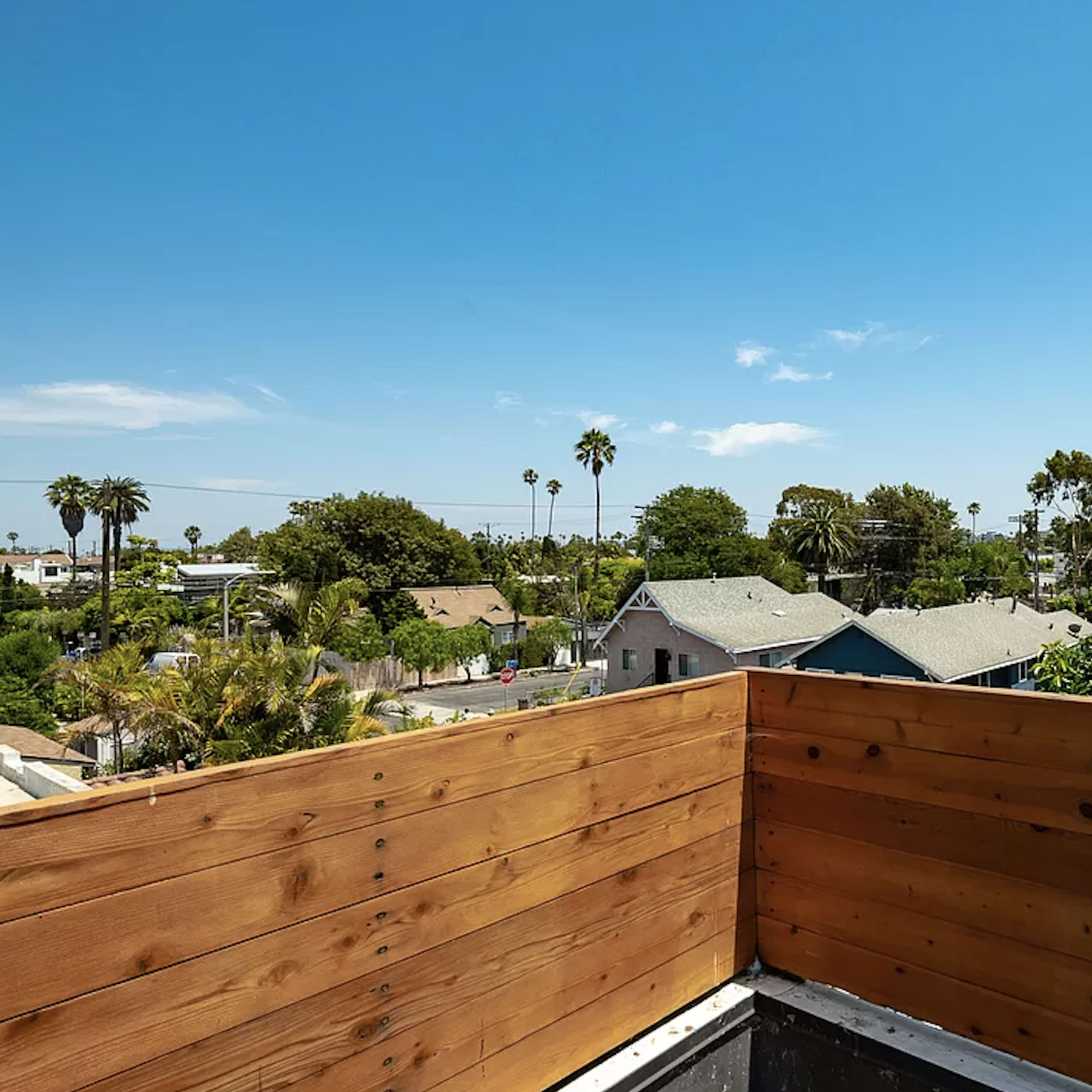 A rooftop view with wooden fencing overlooking a neighborhood lined with palm trees and various houses under a clear blue sky.