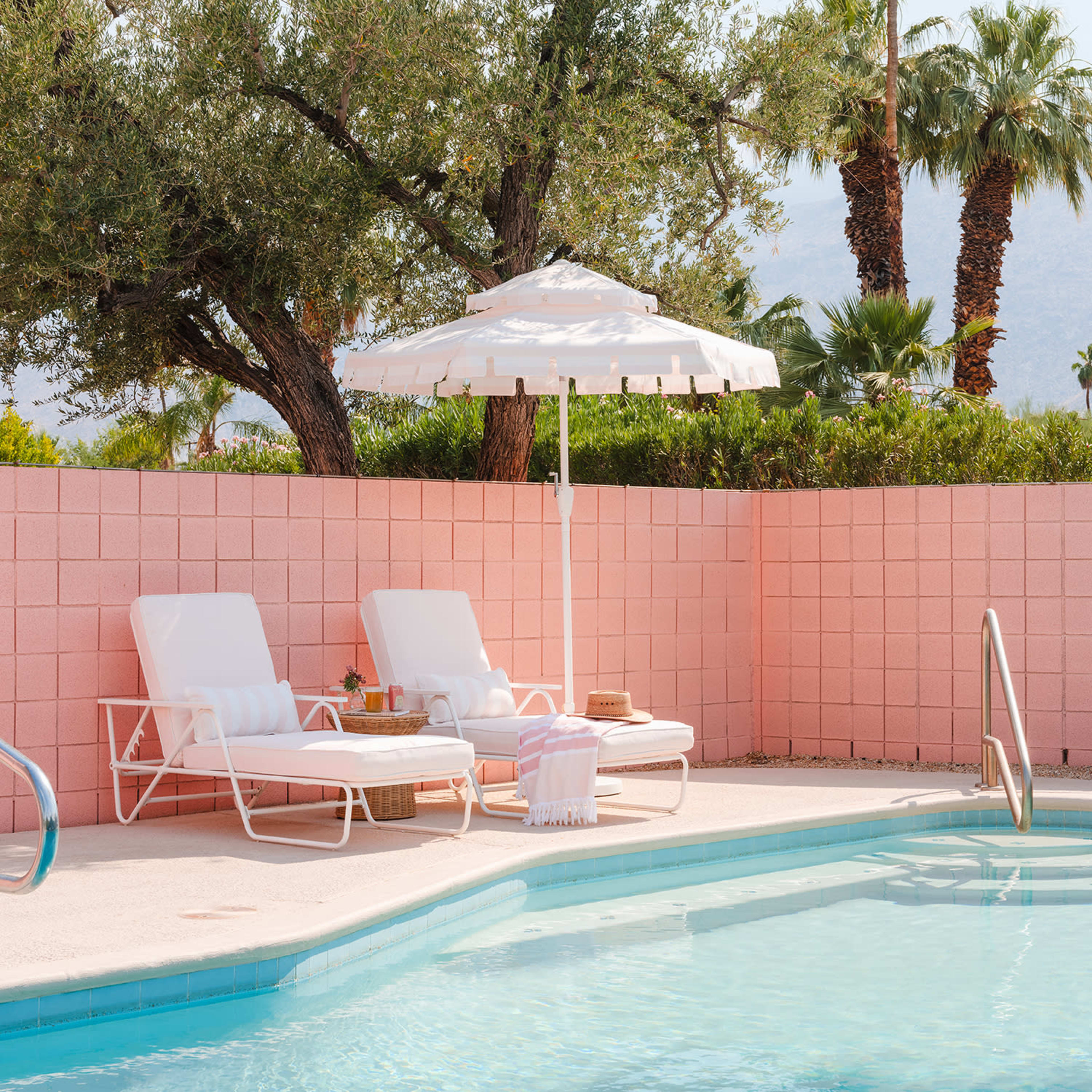 A pink-walled pool area with two lounge chairs, an umbrella, and palm trees in the background.