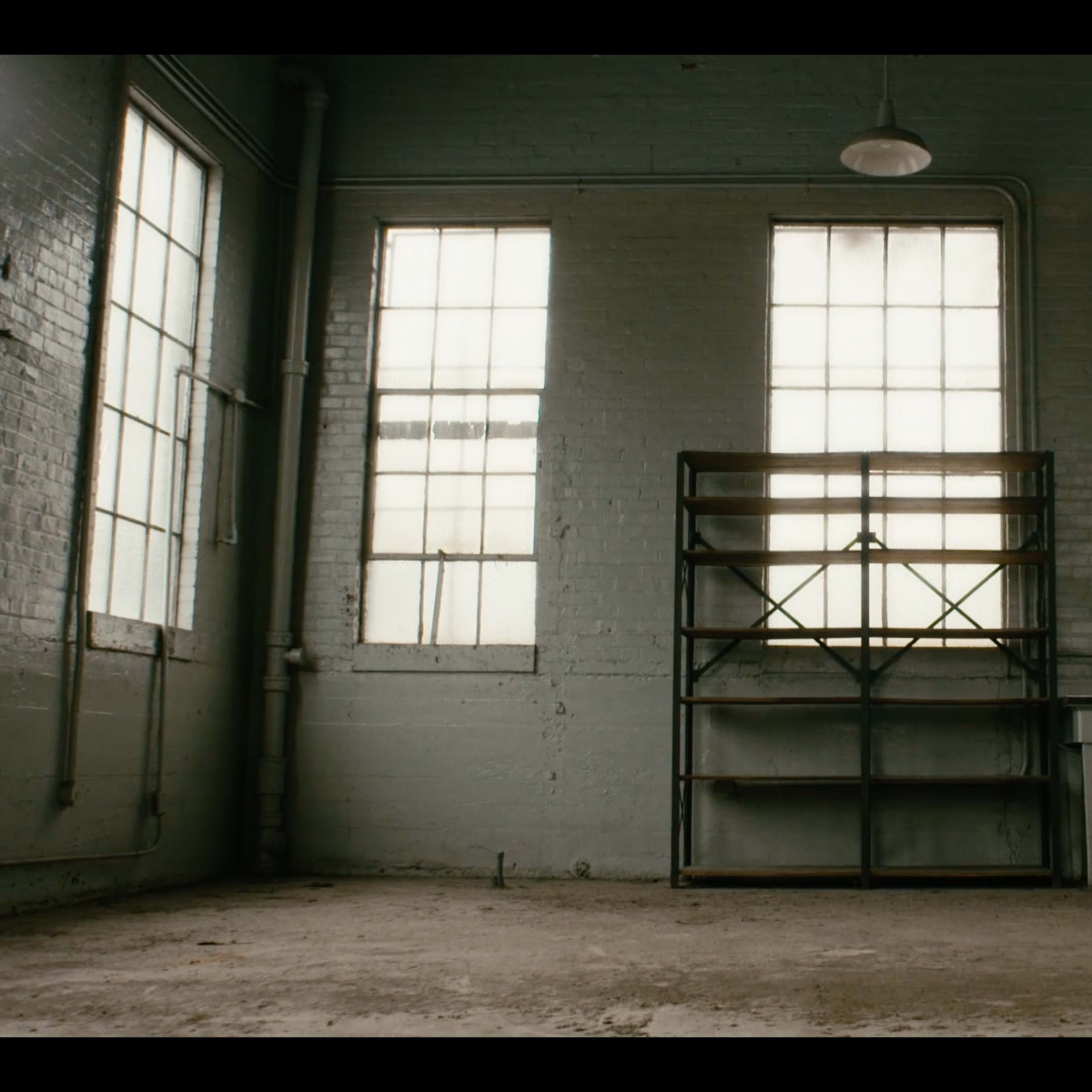An empty, well-lit room with large windows, a metal shelving unit against one wall, and a small table with drawers.