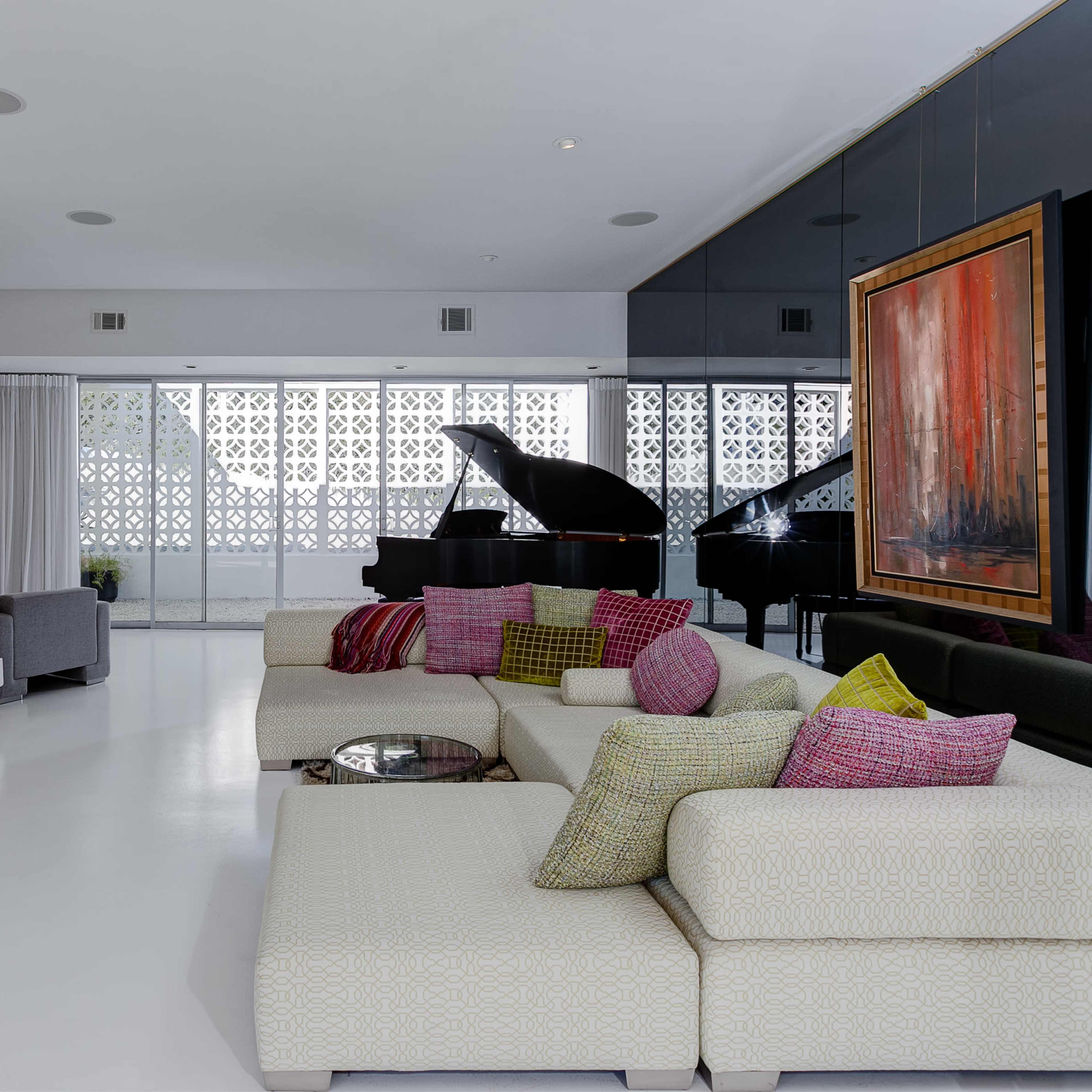 A modern living room with a white floor, sectional sofas adorned with colorful pillows, a wall-mounted television, a grand piano in the background, and large windows with patterned glass.