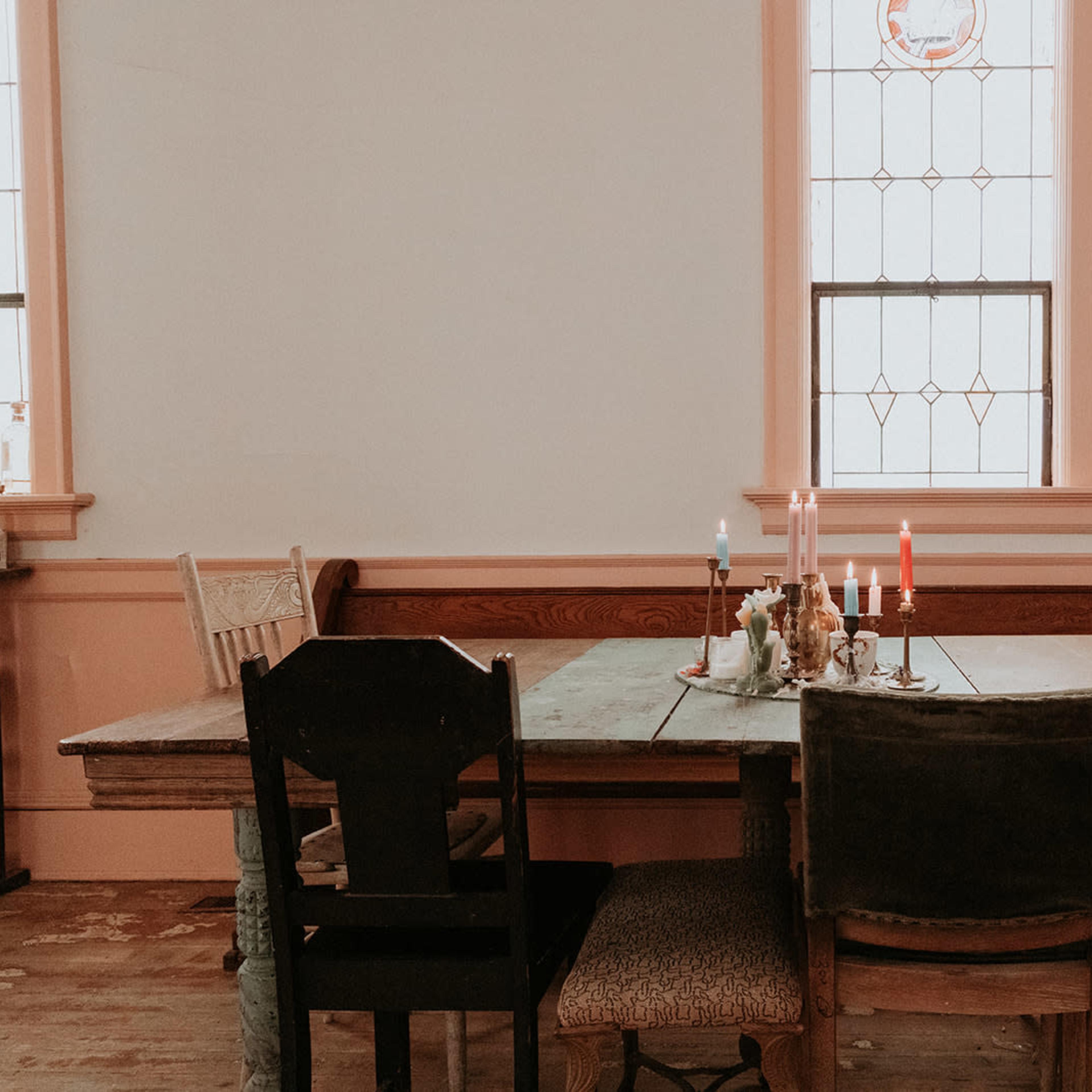 A rustic wooden dining table with various colored candles sits in a room featuring stained glass windows and wooden benches.
