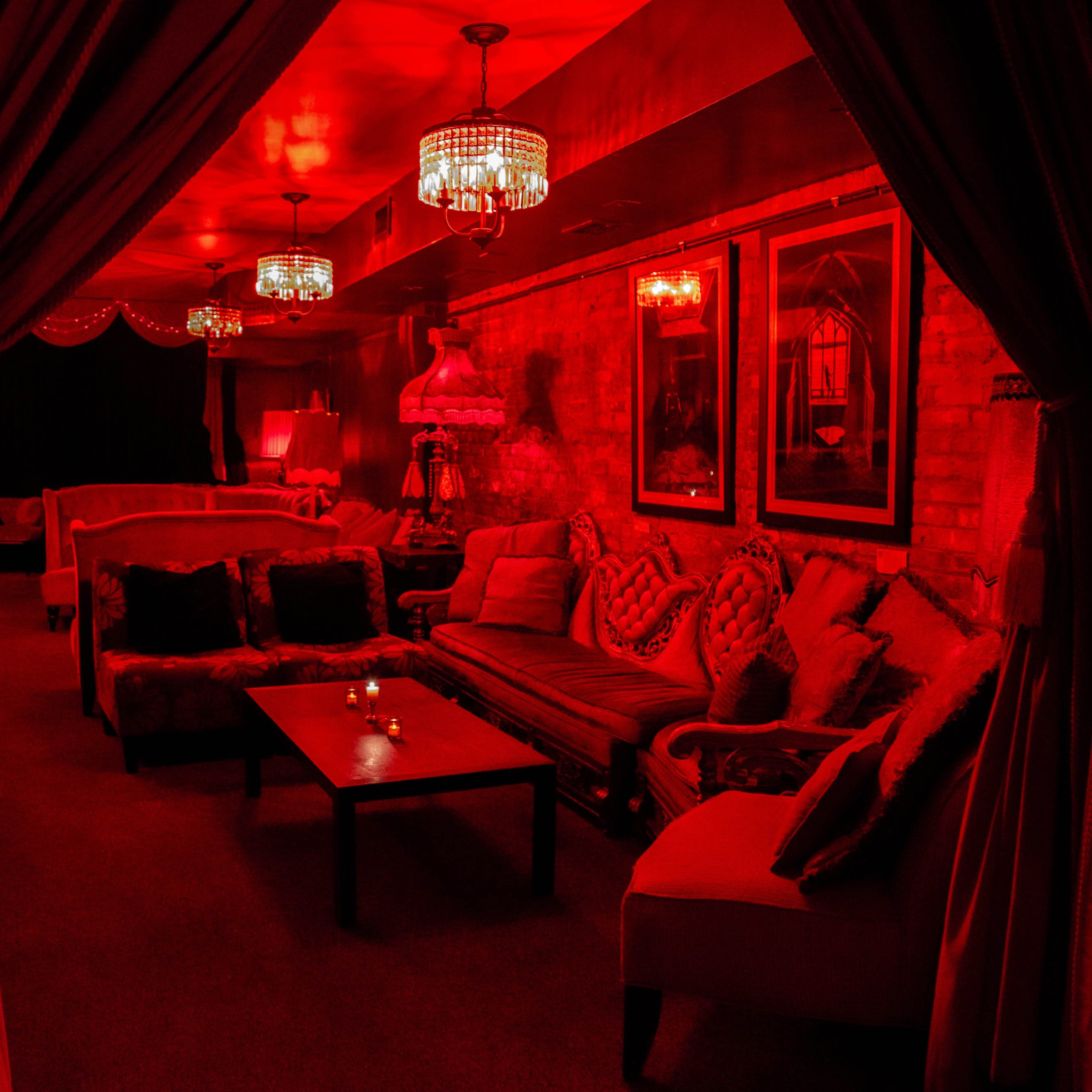 A lounge area is illuminated with red light, featuring plush seating and decorative chandeliers.