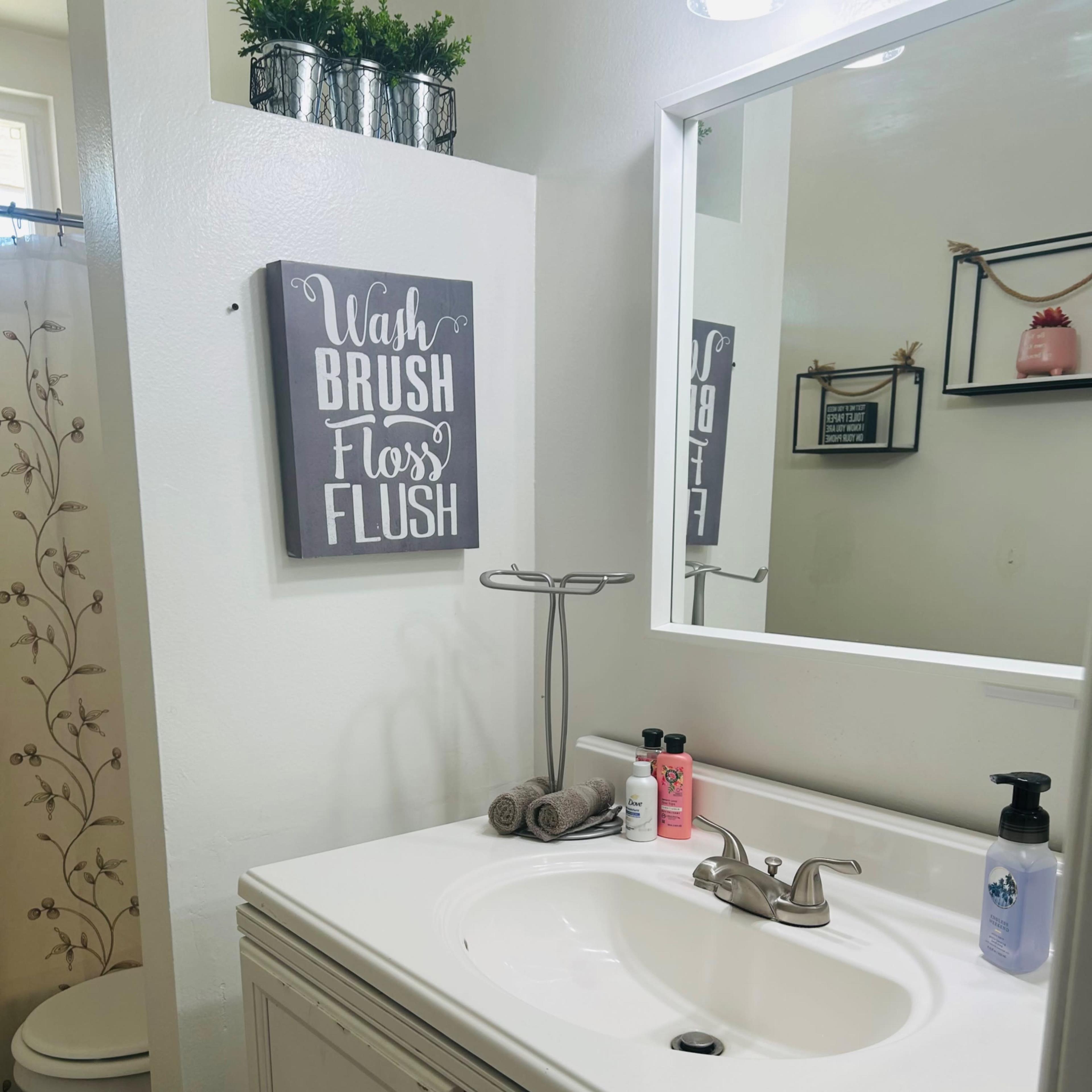 A compact bathroom featuring a white vanity with a sink, a framed wall sign, and a toilet in the background.