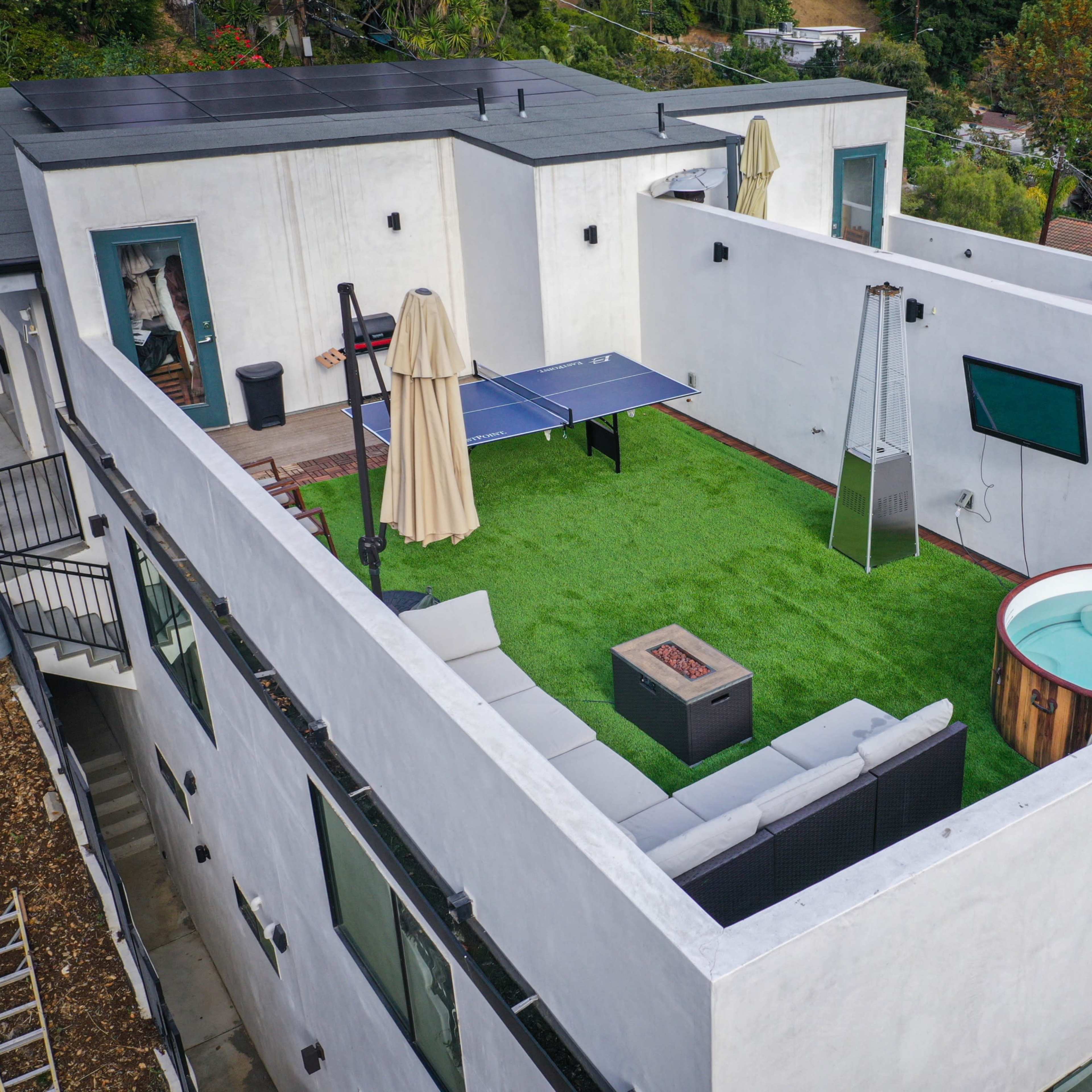 A rooftop terrace featuring a green artificial lawn, a ping pong table, a small hot tub, a television mounted on a wall, and a seating area with modern furniture.
