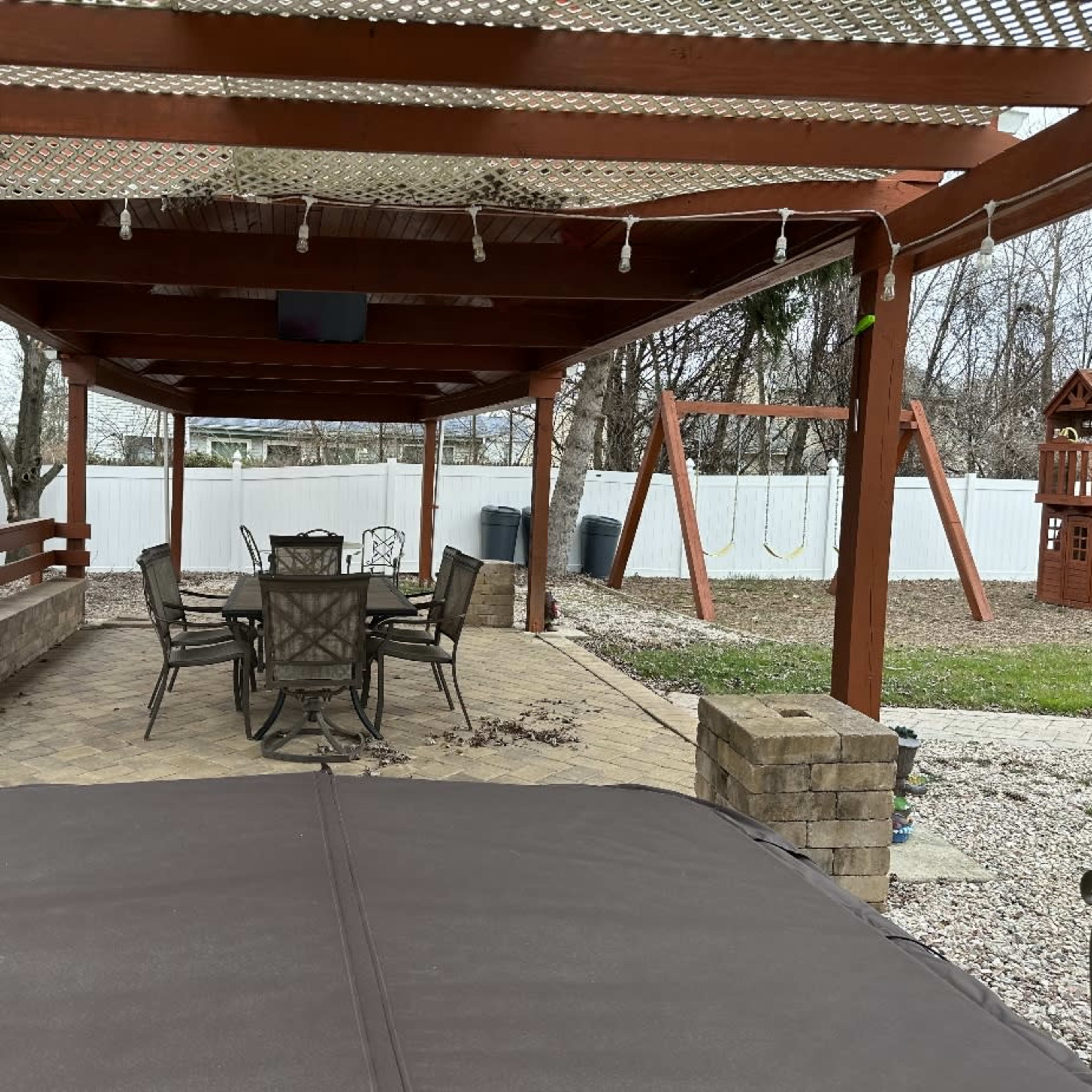 A covered patio area with a dining table and chairs, surrounded by a stone wall and featuring swings in the background.