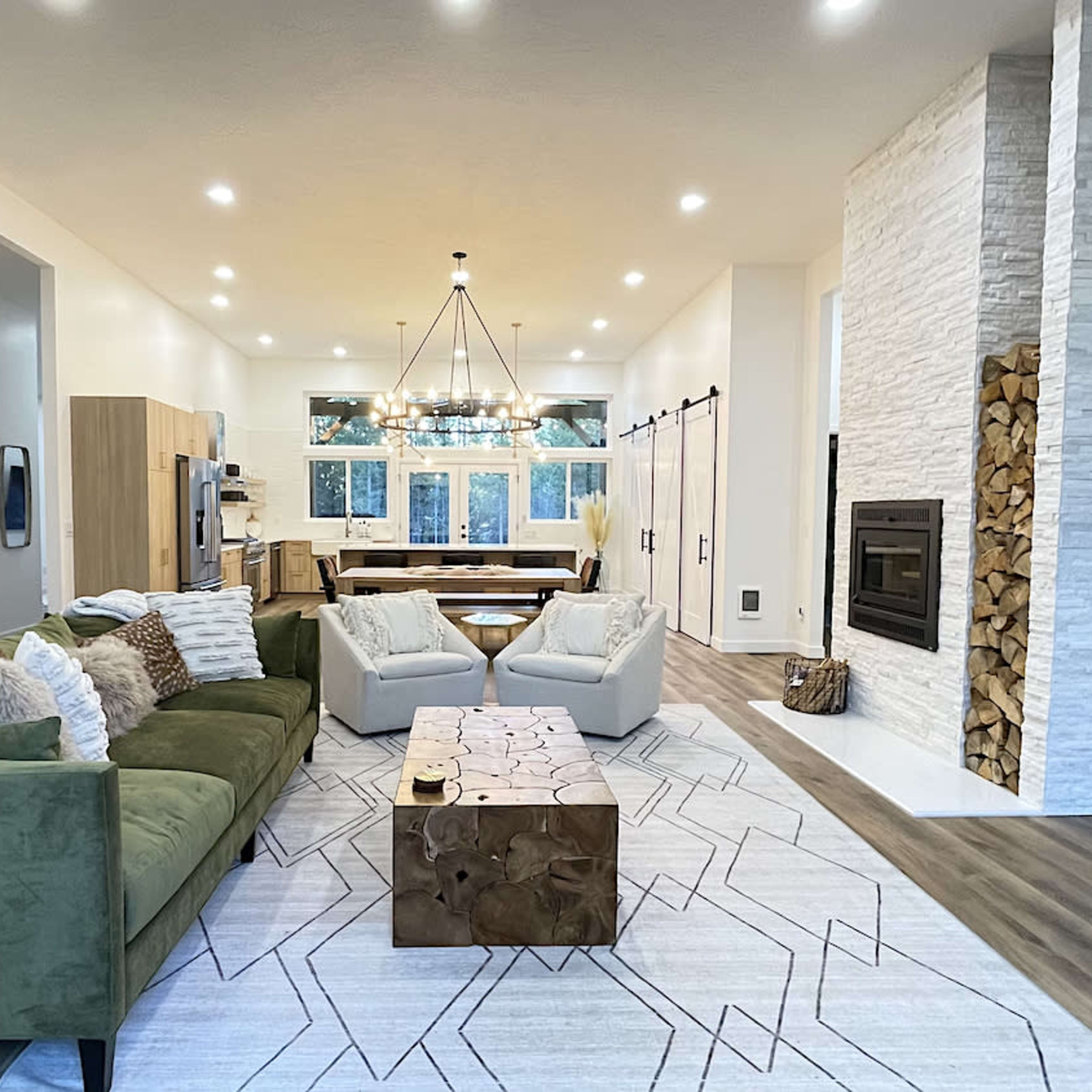 The living room features a green sofa, two armchairs, a wooden coffee table, and a fireplace surrounded by stacked wood, all arranged under a chandelier with large windows in the background.