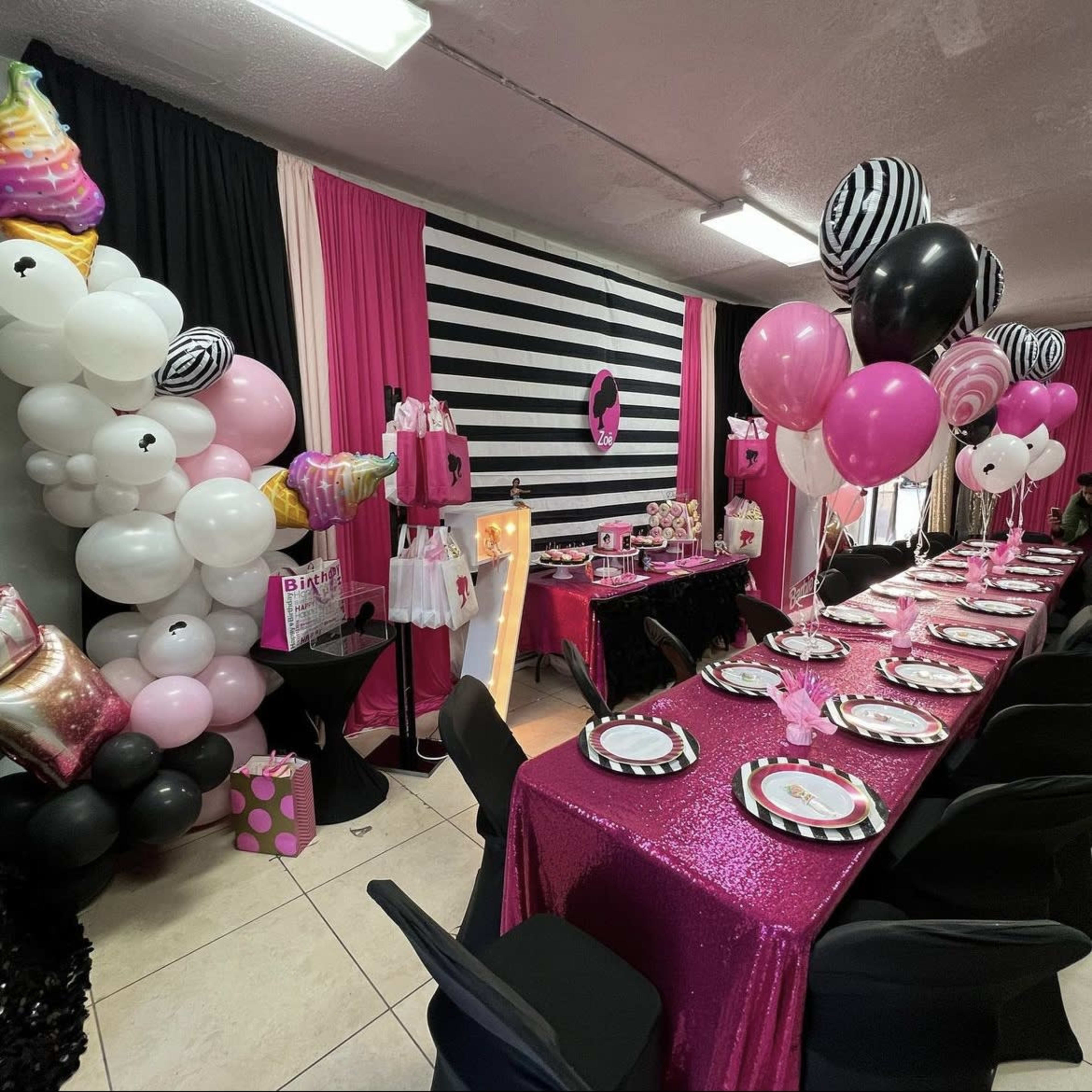 A decorated party space featuring a long table covered with a sequined pink tablecloth, surrounded by balloons and themed decorations in black and pink.