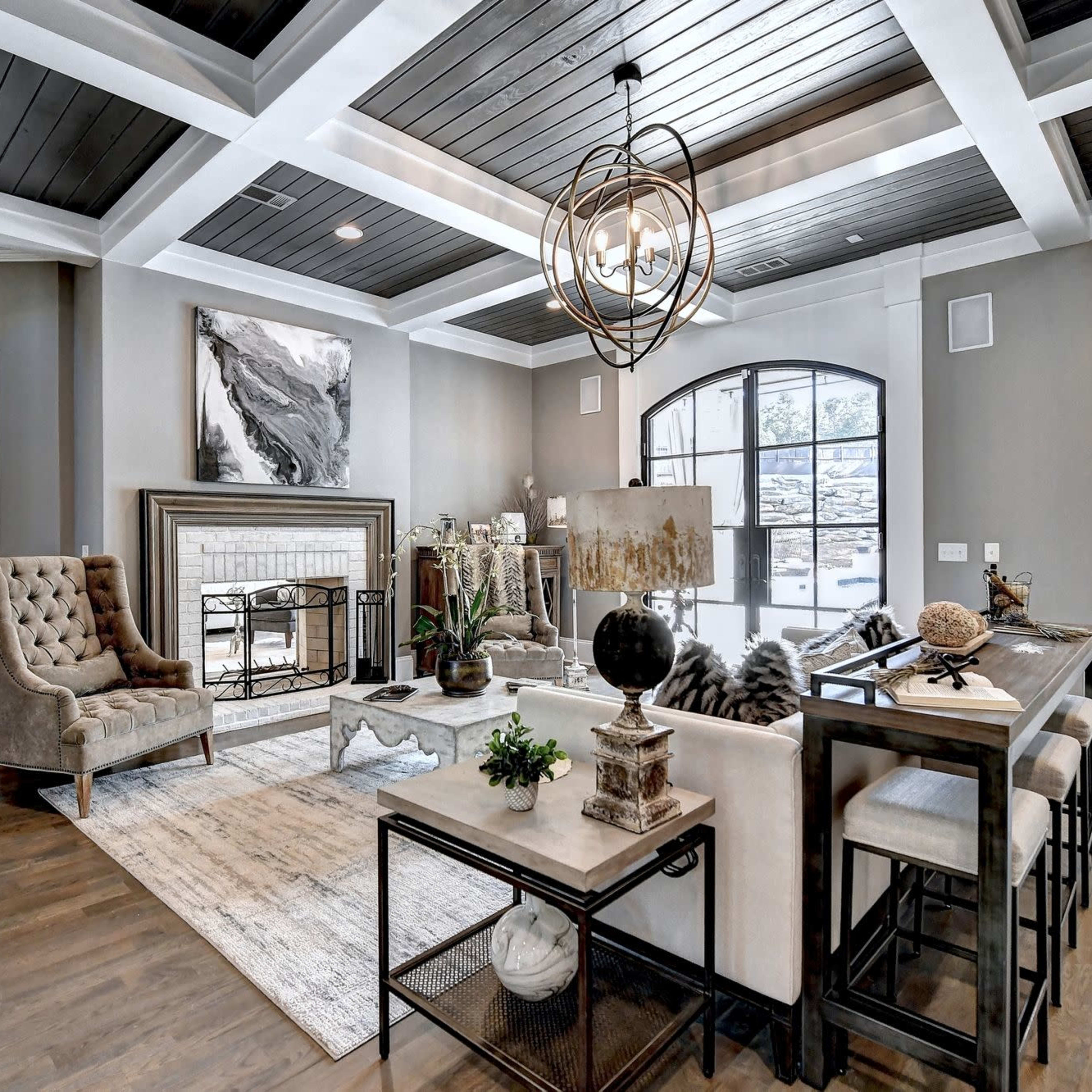 A spacious living room featuring a mix of modern and traditional furnishings, including a tufted chair, a wooden dining table, and a fireplace, all under a coffered ceiling.
