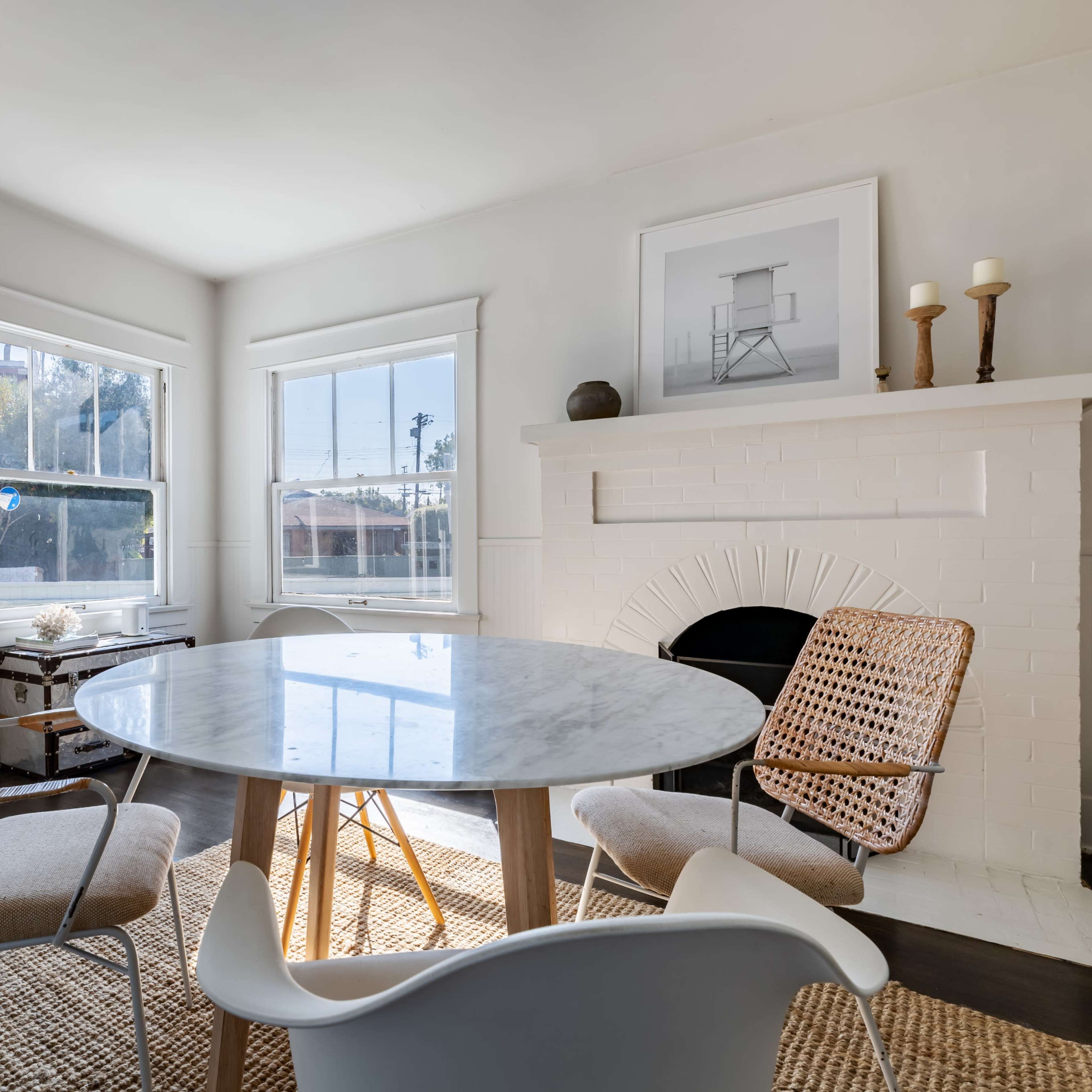 A bright dining area features a round marble table surrounded by woven chairs and a white brick fireplace with candles.